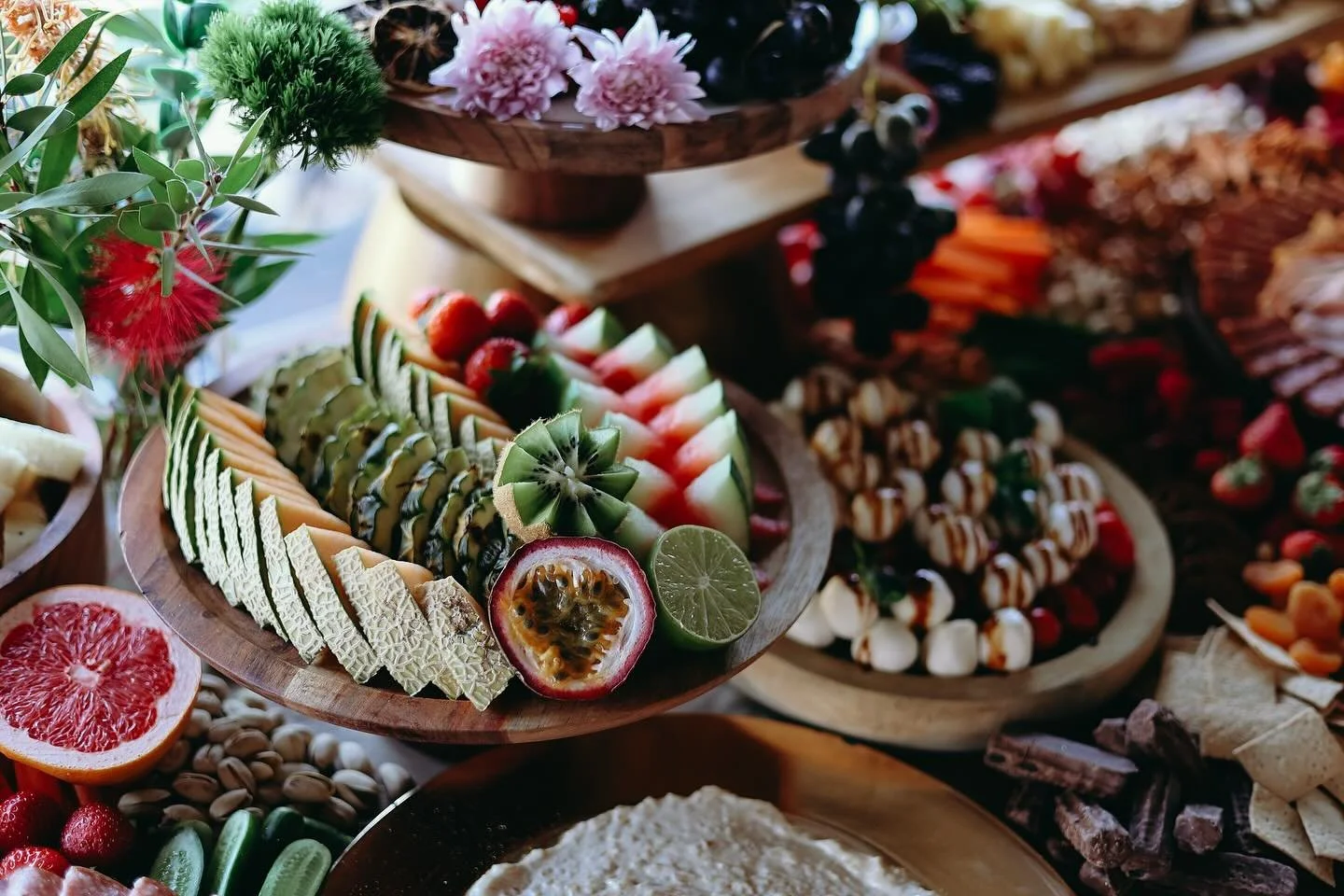 A colorful display of assorted fruits, cheeses, and snacks on wooden platters, including slices of watermelon, pineapple, passion fruit, lime, strawberries, and a variety of candies and chocolates.