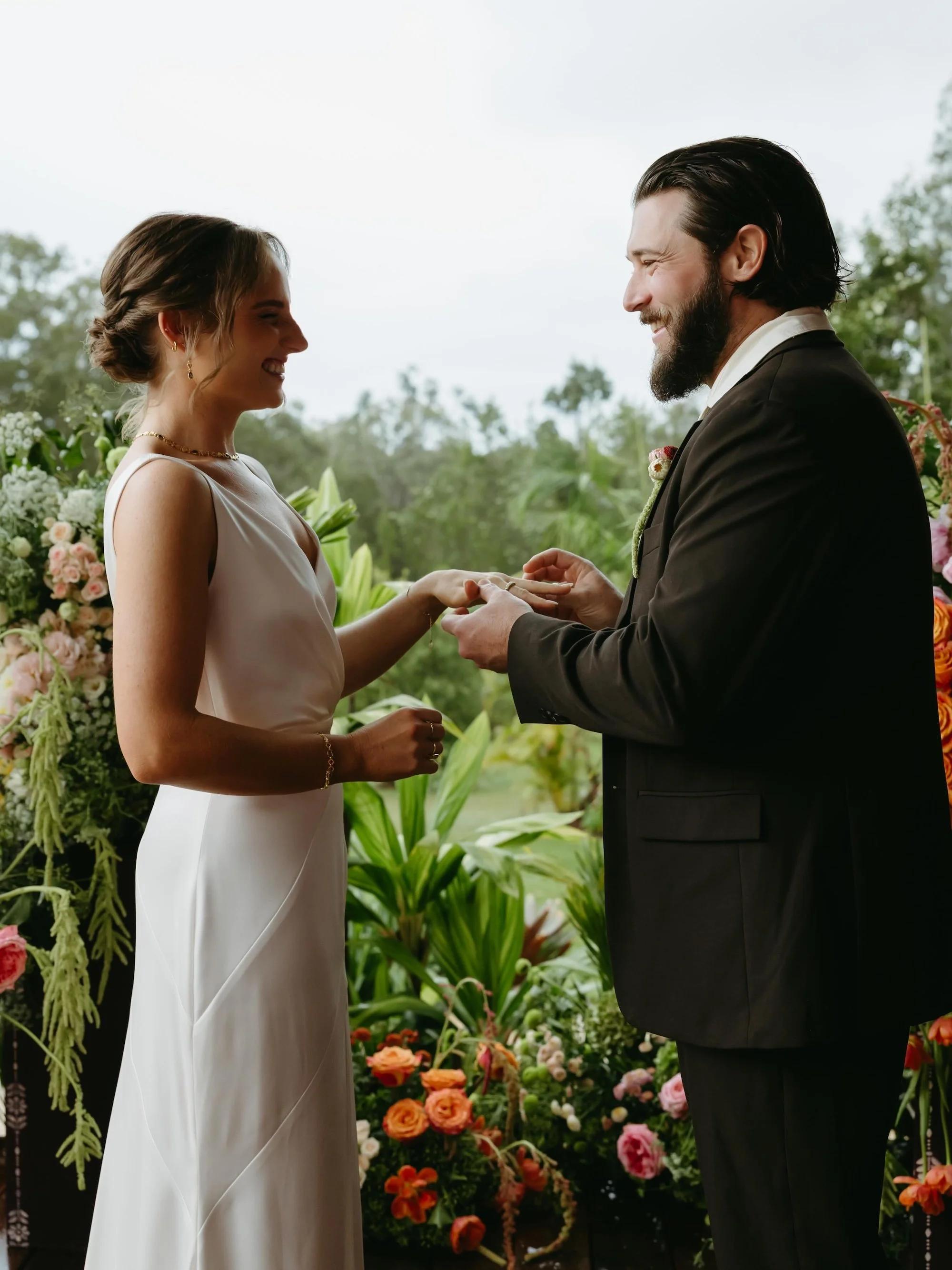 A bride and groom exchanging vows at an outdoor wedding ceremony, standing in front of a colorful floral backdrop with greenery and trees in the background, both smiling. | Goolawah Retreat Crescent Head