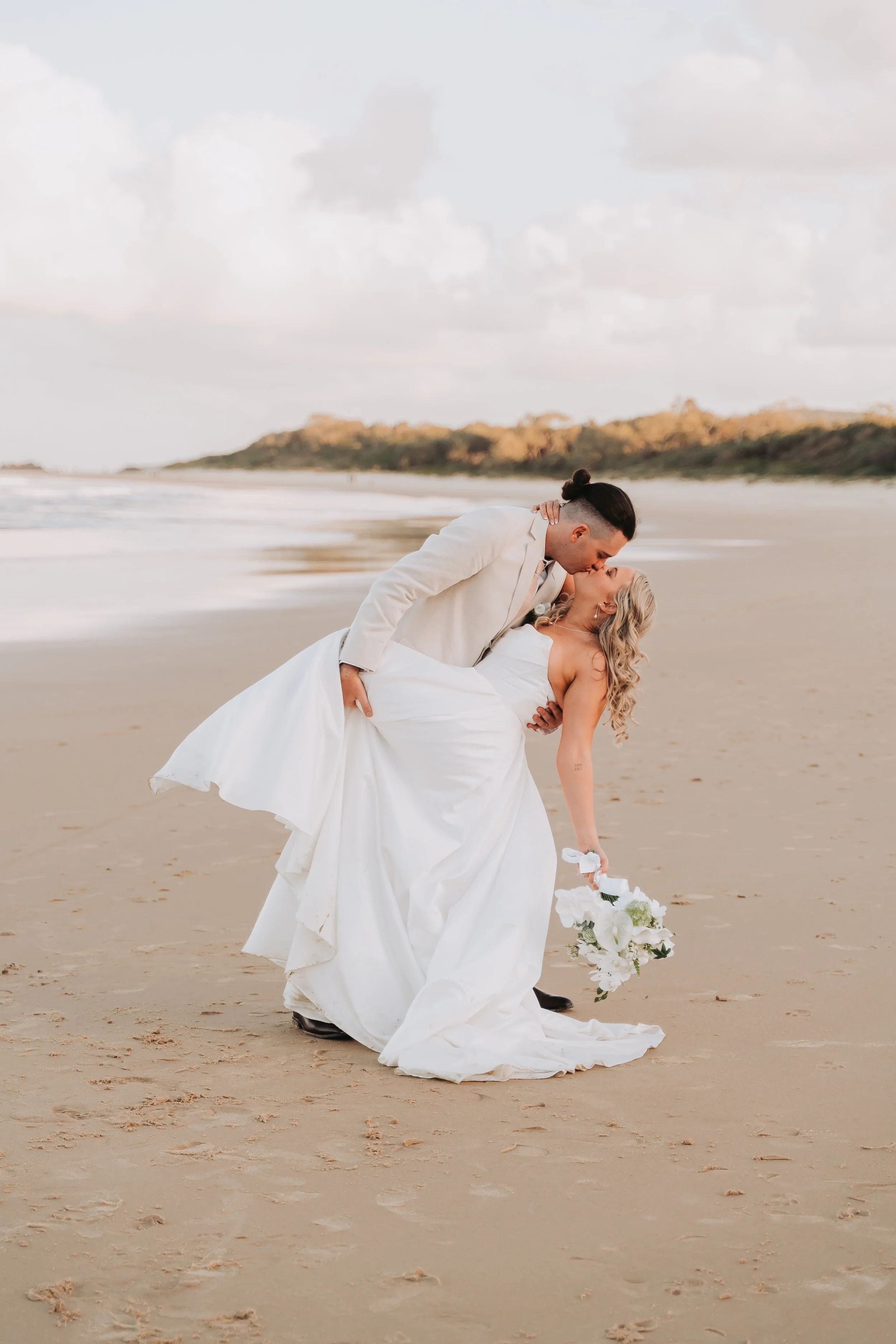A bride and groom on a sandy beach, with the groom dipping the bride as she holds a bouquet of white flowers, both in wedding attire, against a backdrop of ocean and cloudy sky.