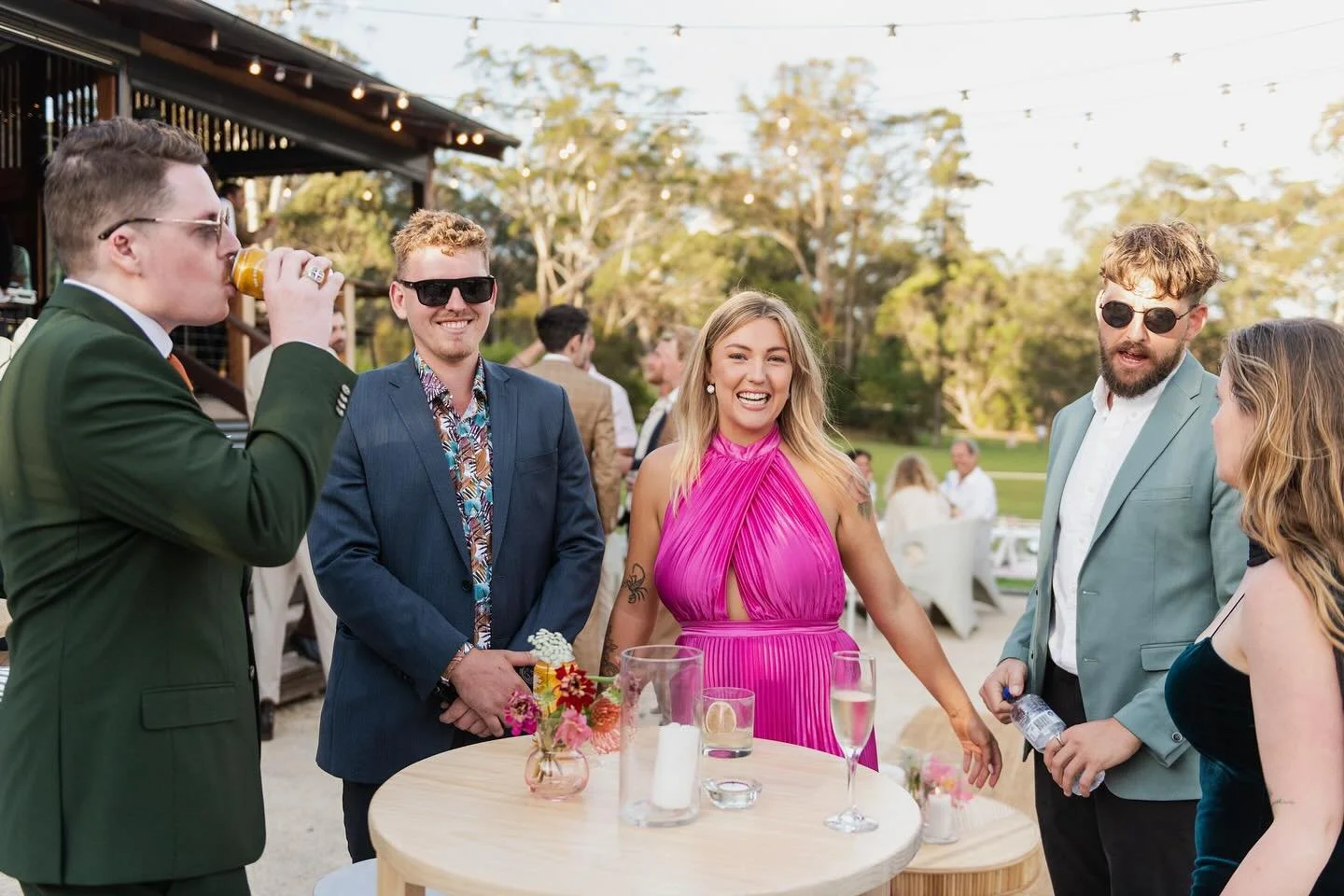 Group of friends at an outdoor party, smiling and chatting around a table with drinks, flowers, and candles, during sunset.