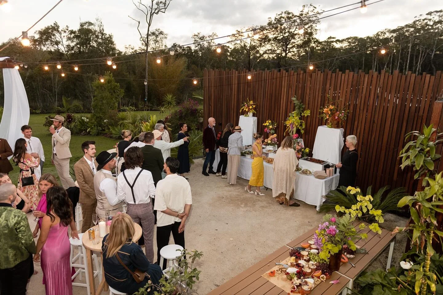 People at an outdoor celebration with string lights, buffet tables with flowers, and a garden backdrop.