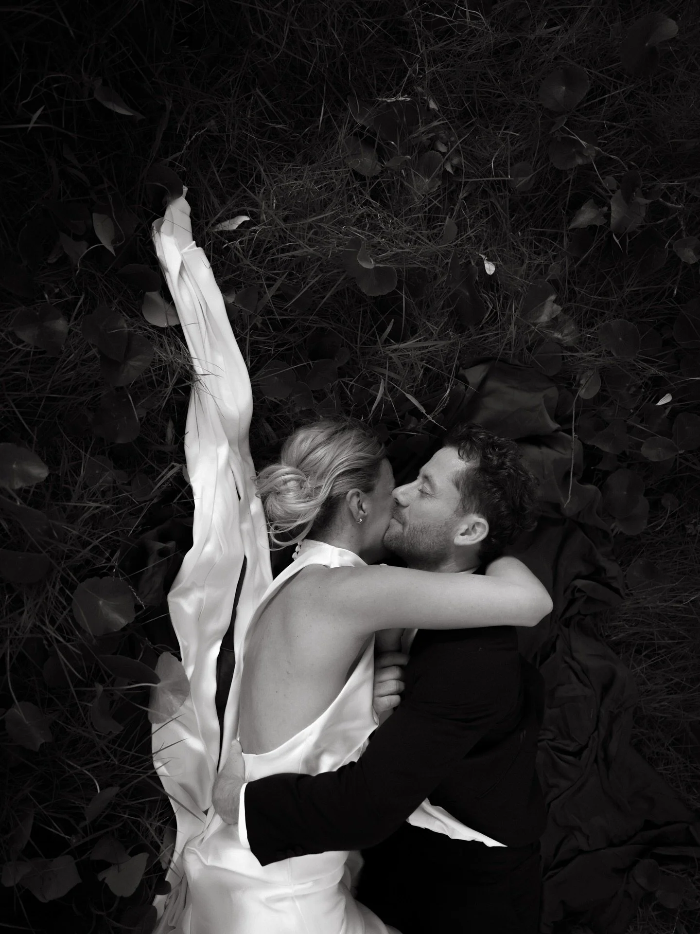 A black and white photo of a couple lying on grass and leaves, kissing, with one woman wearing a white dress and the man in dark clothing.