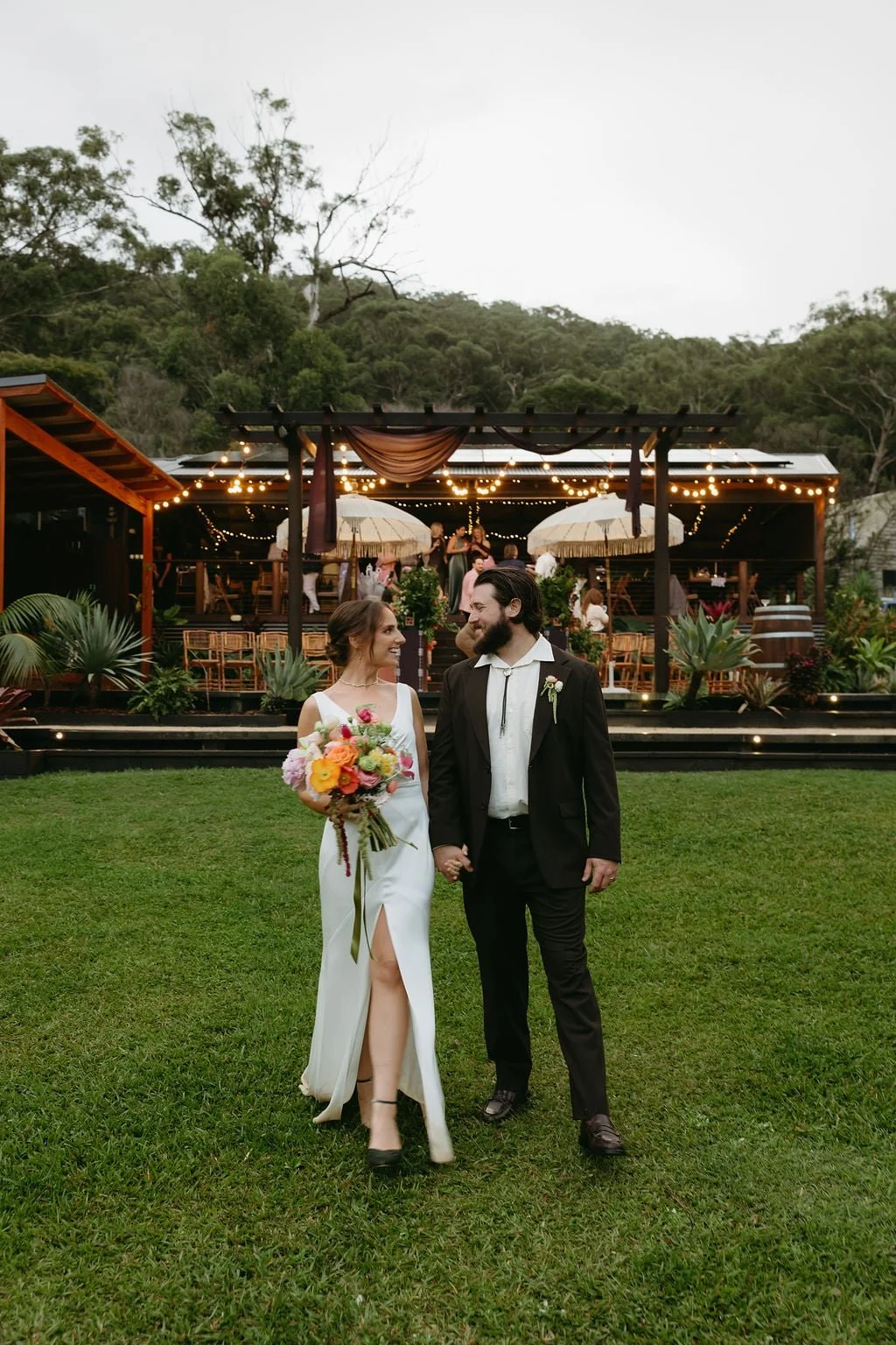 A bride and groom holding hands and smiling at each other on a grassy lawn, with a decorated outdoor wedding reception area featuring string lights, umbrellas, and guests in the background, surrounded by trees.