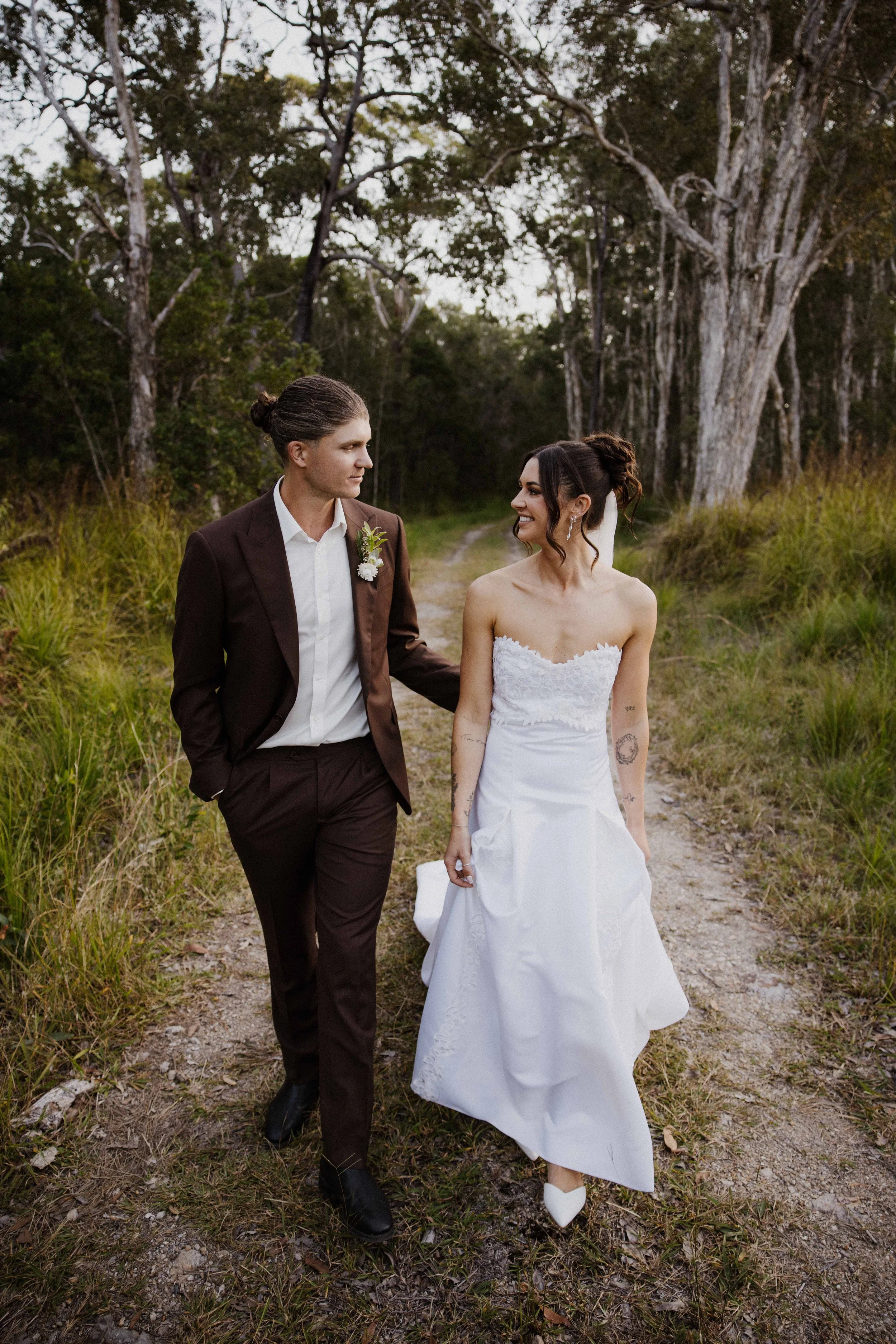 A couple dressed in wedding attire walking along a forest trail. The groom is wearing a dark suit and the bride is in a strapless white wedding gown. They are holding hands and looking at each other with smiles. Trees and grass surround the path.