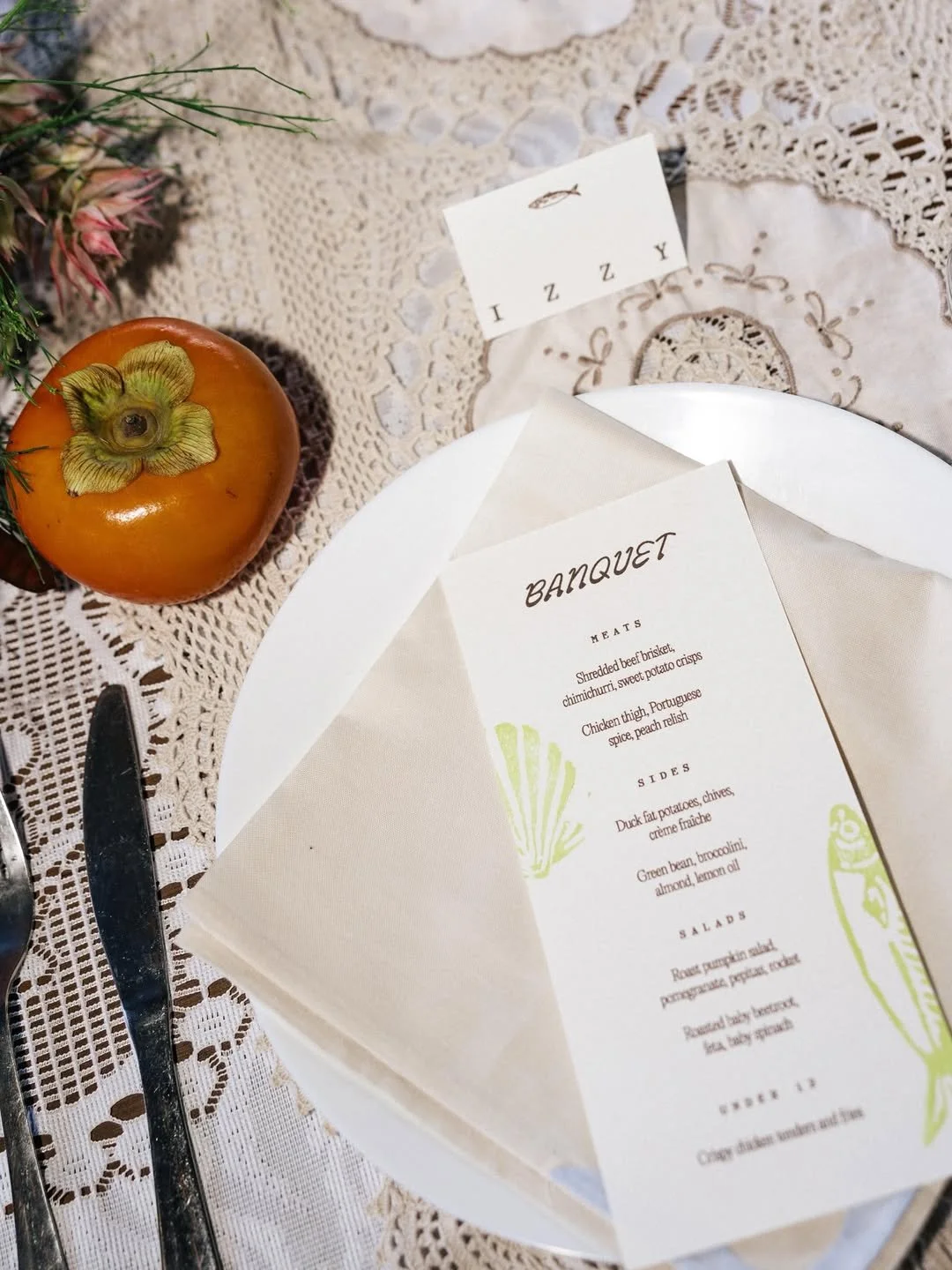 A banquet menu on a white plate, designed with green illustrations, placed on a linen tablecloth with lace details. Beside the plate, there is a persimmon fruit, and some greenery, along with a small white place card.