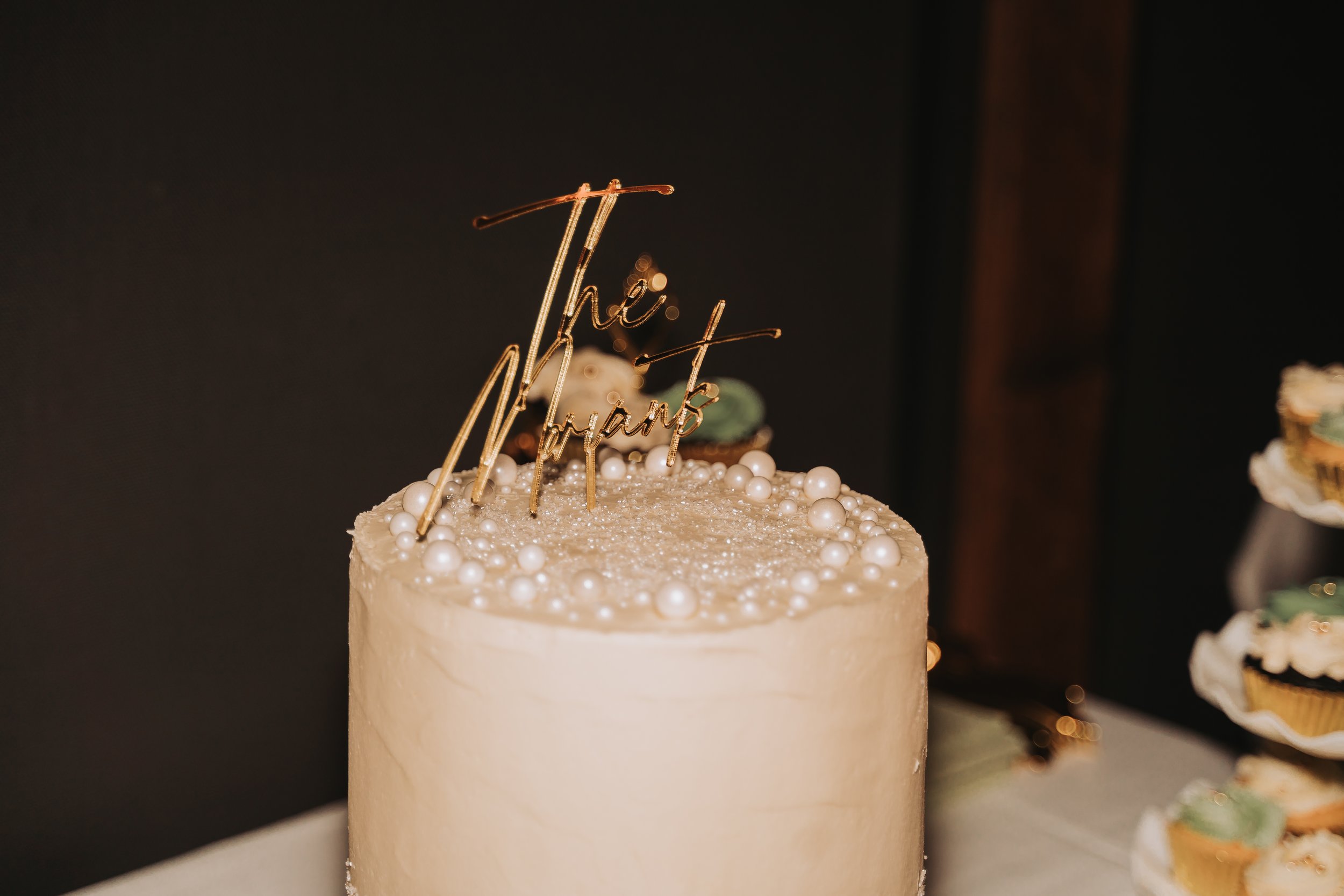 A white frosted birthday cake decorated with white pearls and a rose. A gold wire cake topper reads 'the best' in cursive script.