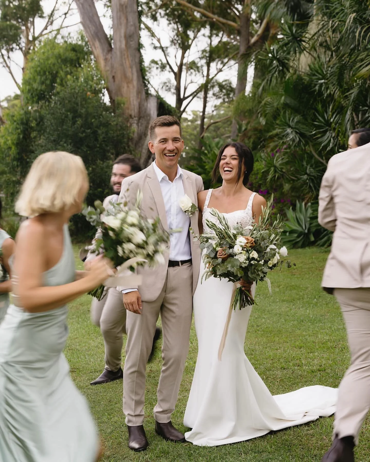 A newlywed couple standing outdoors among friends, smiling and holding bouquets of flowers, during a wedding celebration in a lush green garden.| Goolawah Retreat Wedding Crescent Head