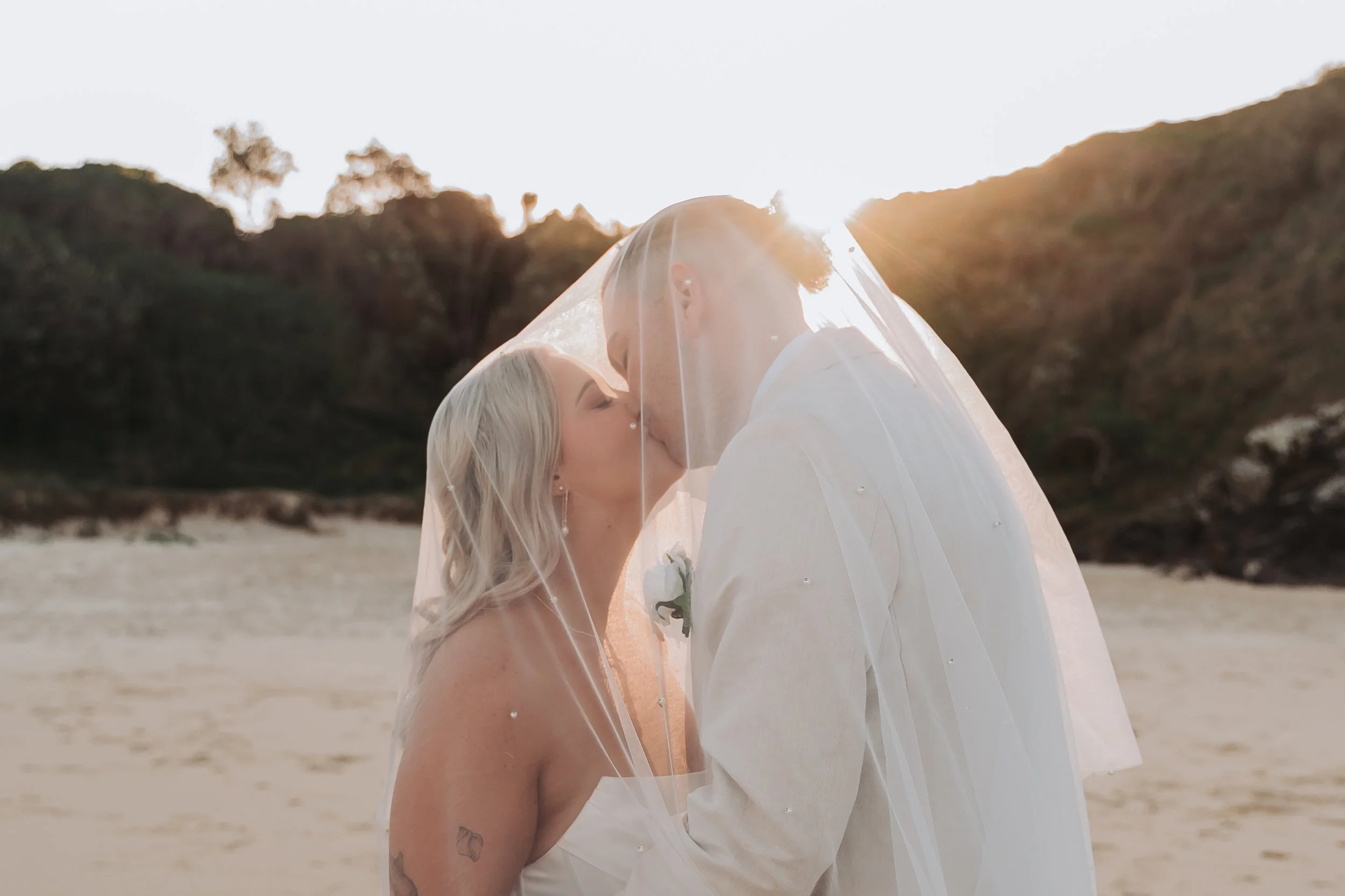 A bride and groom sharing a kiss on a beach at sunset, with the bride in a white dress and veil, and the groom in a white suit.