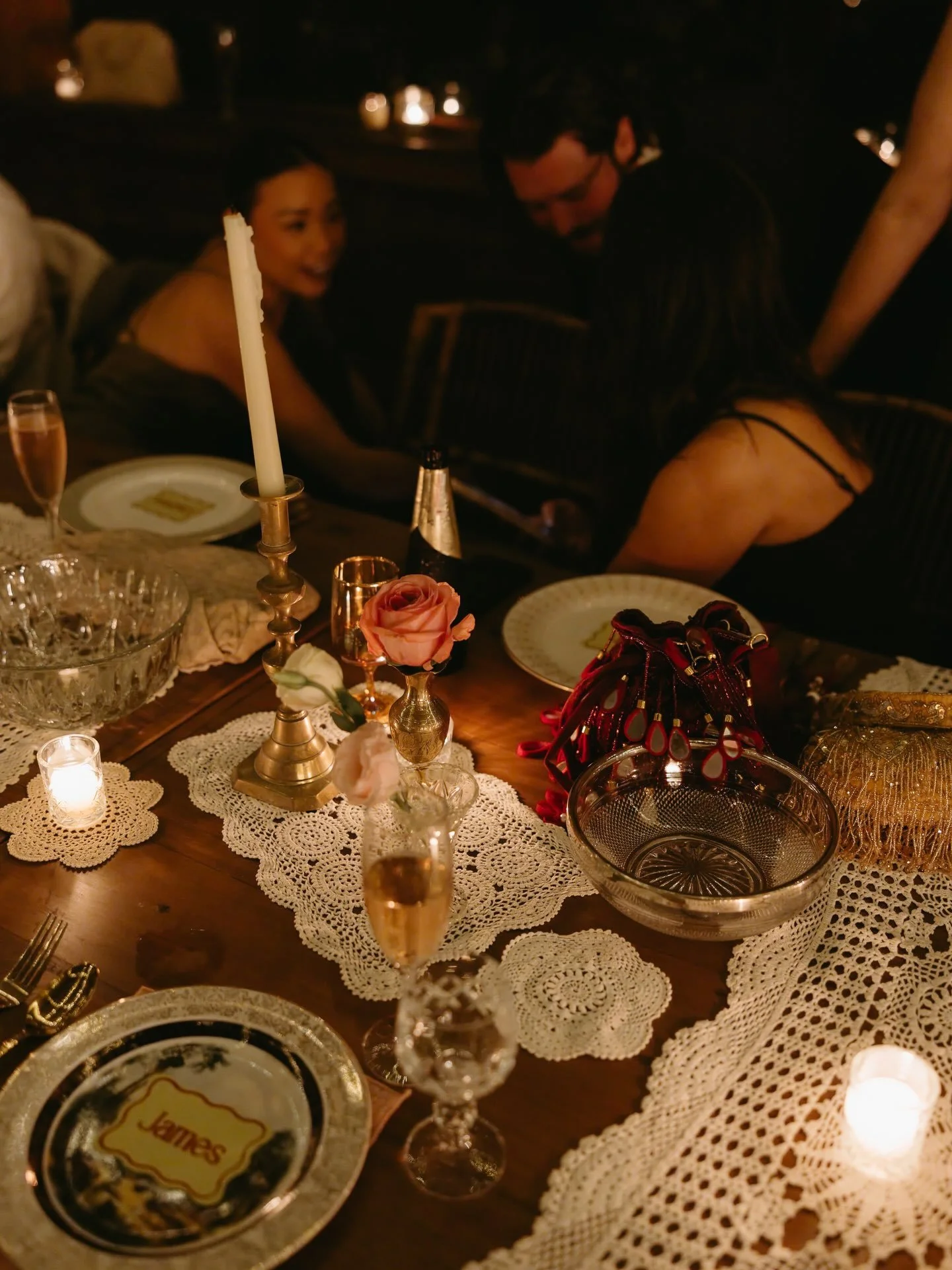 A table set for a celebration with vintage dishes, candles, and drinks. In the background, people are socializing in a dimly lit room.