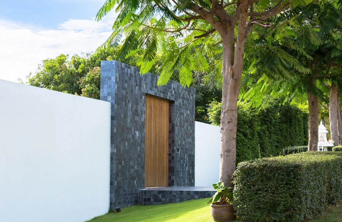 Modern wooden door set in dark stone wall with white plaster extension, surrounded by green grass, trees, and bushes.