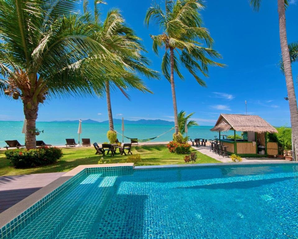 Tropical beach scene with a swimming pool, palm trees, sun loungers, and a thatched-roof bar overlooking the ocean under a clear blue sky.