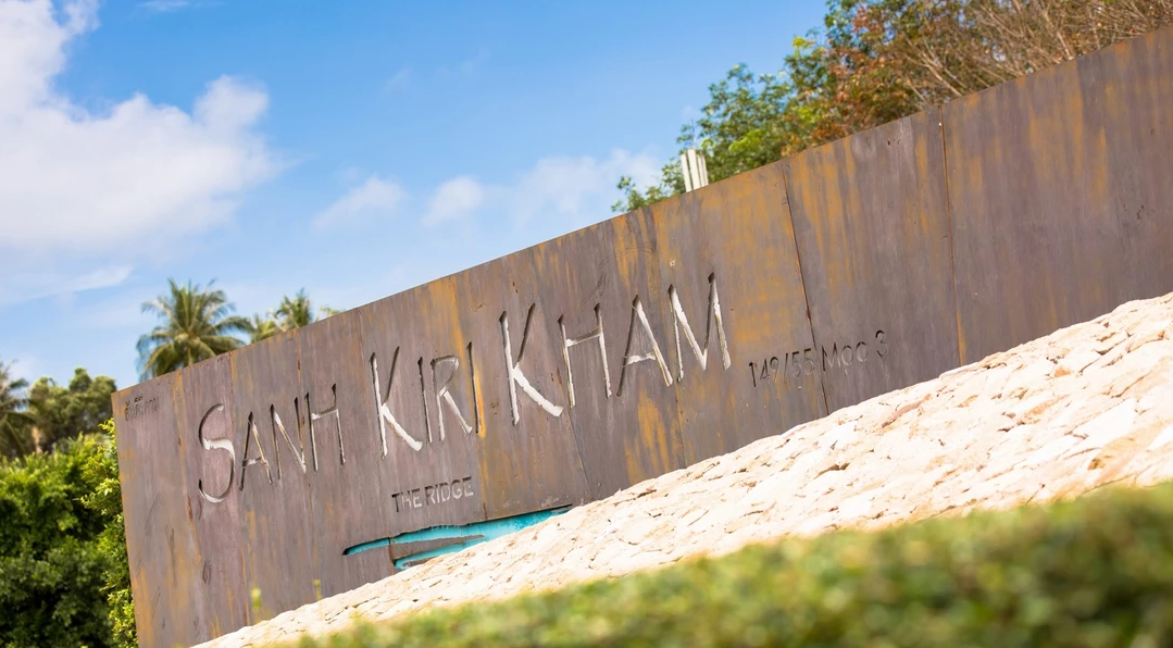 Rustic metal sign with "Sanh Kiri Kham" and "The Ridge" engraving, surrounded by greenery and palm trees, against a blue sky.