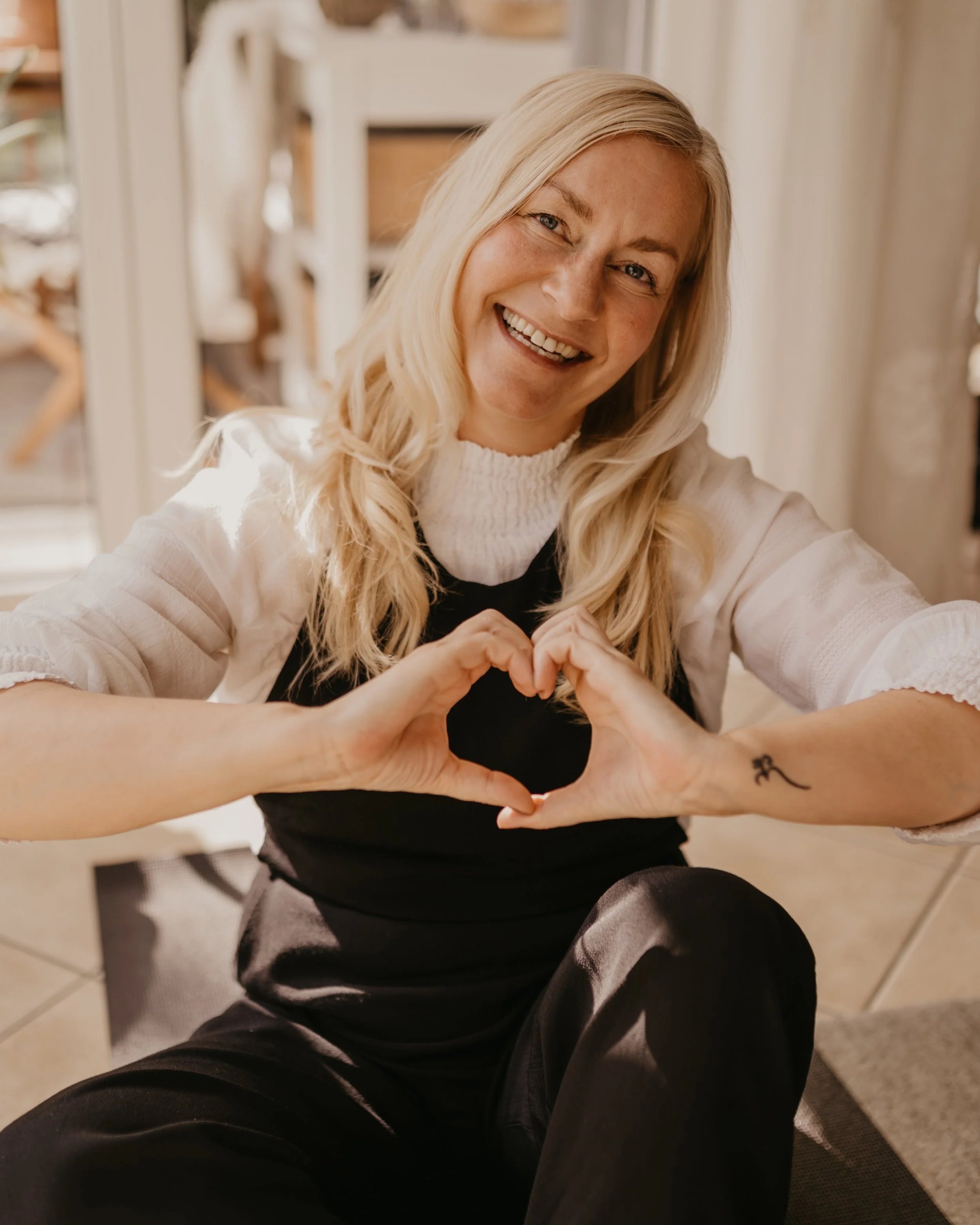 Yvonne sitzt am Boden und formt beide Hände zu einem Herz und lächelt.