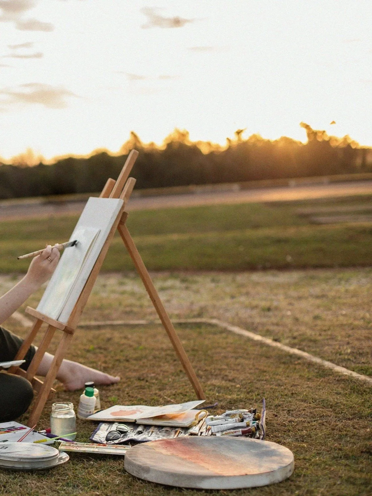Frau sitzt vor Staffelei in Grünen und malt, neben ihr Pinsel, Farbtuben, Wasserglas ein Malbuch