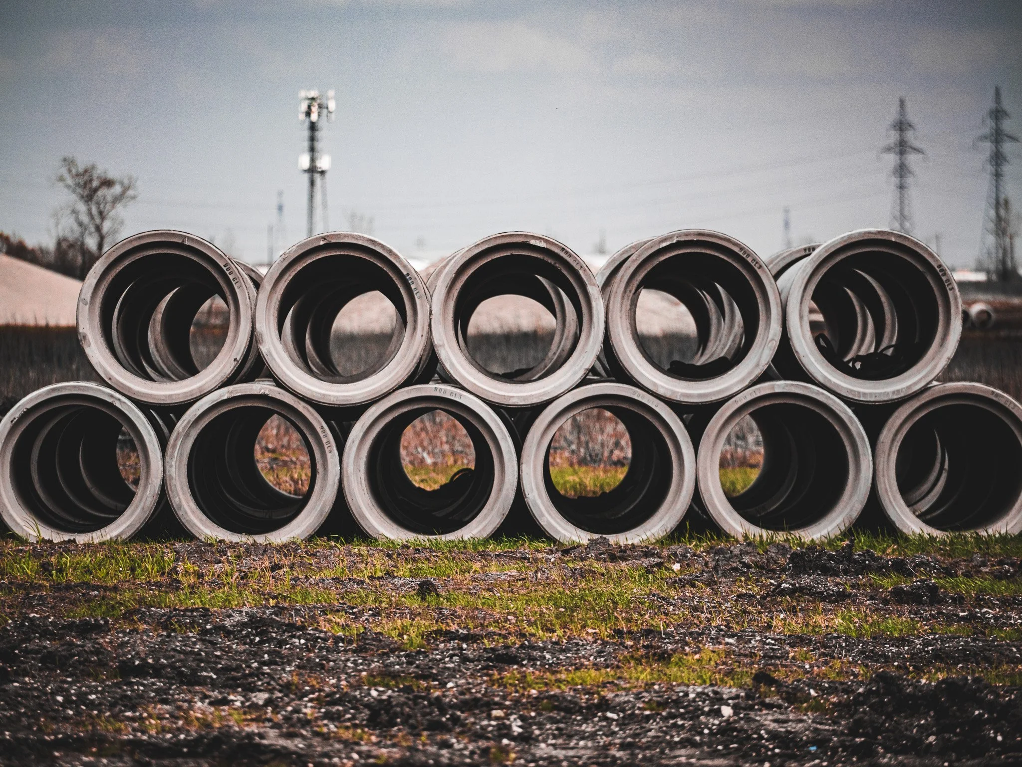 A stack of large gray concrete pipes horizontally arranged outdoors on grassy and gravel ground with power lines and a cloudy sky in the background.