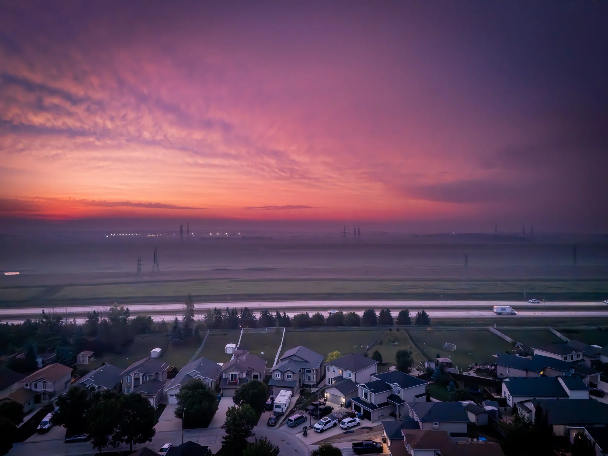 Sunset over a suburban neighborhood with houses, streets, and trees in the foreground, and a cloudy sky with shades of pink, purple, and orange in the background Winnipeg Manitoba.