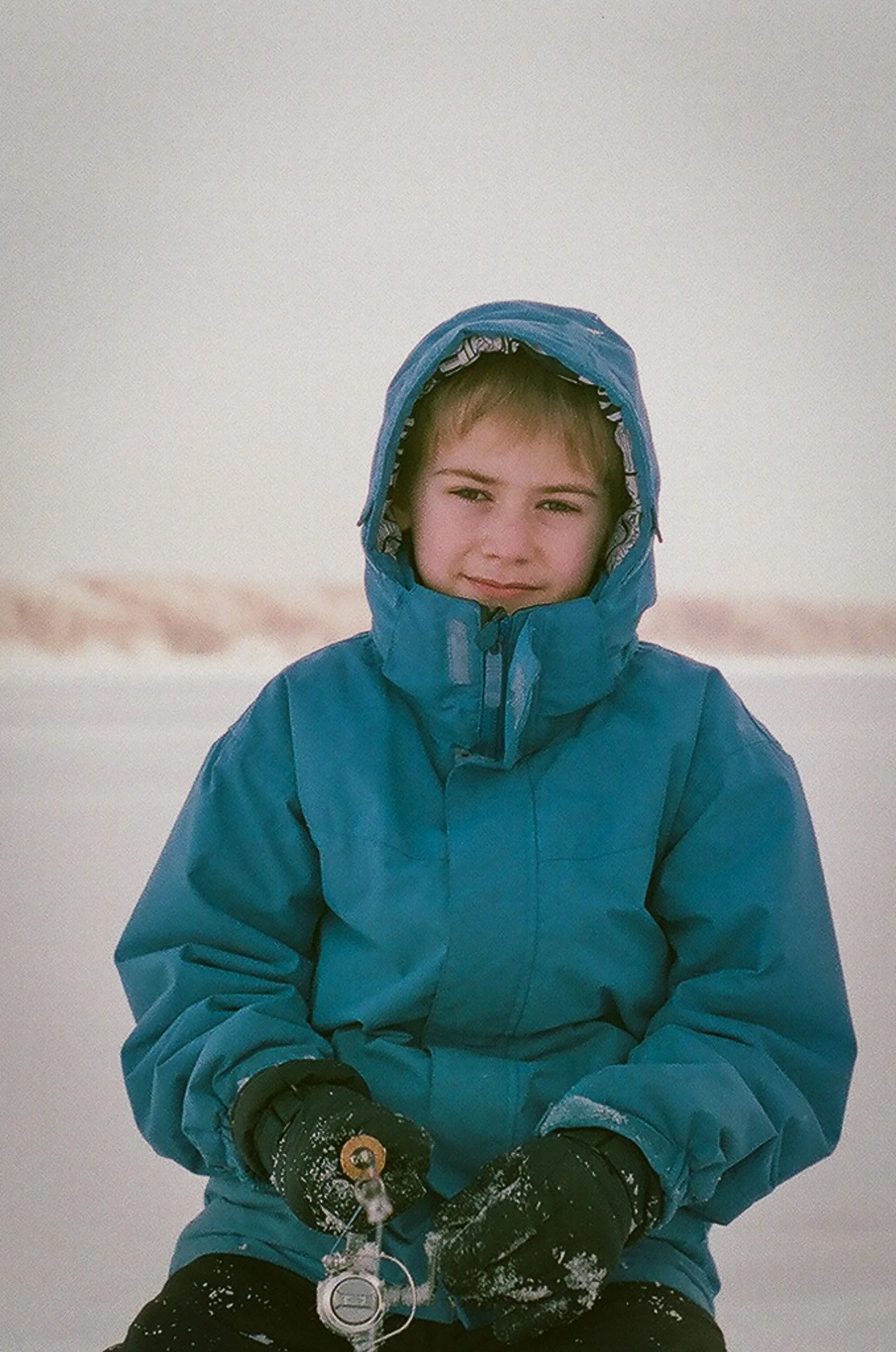 A young person outdoors in winter clothing, wearing a blue jacket with the hood up, black gloves, and holding snow and ice, with a snowy background.