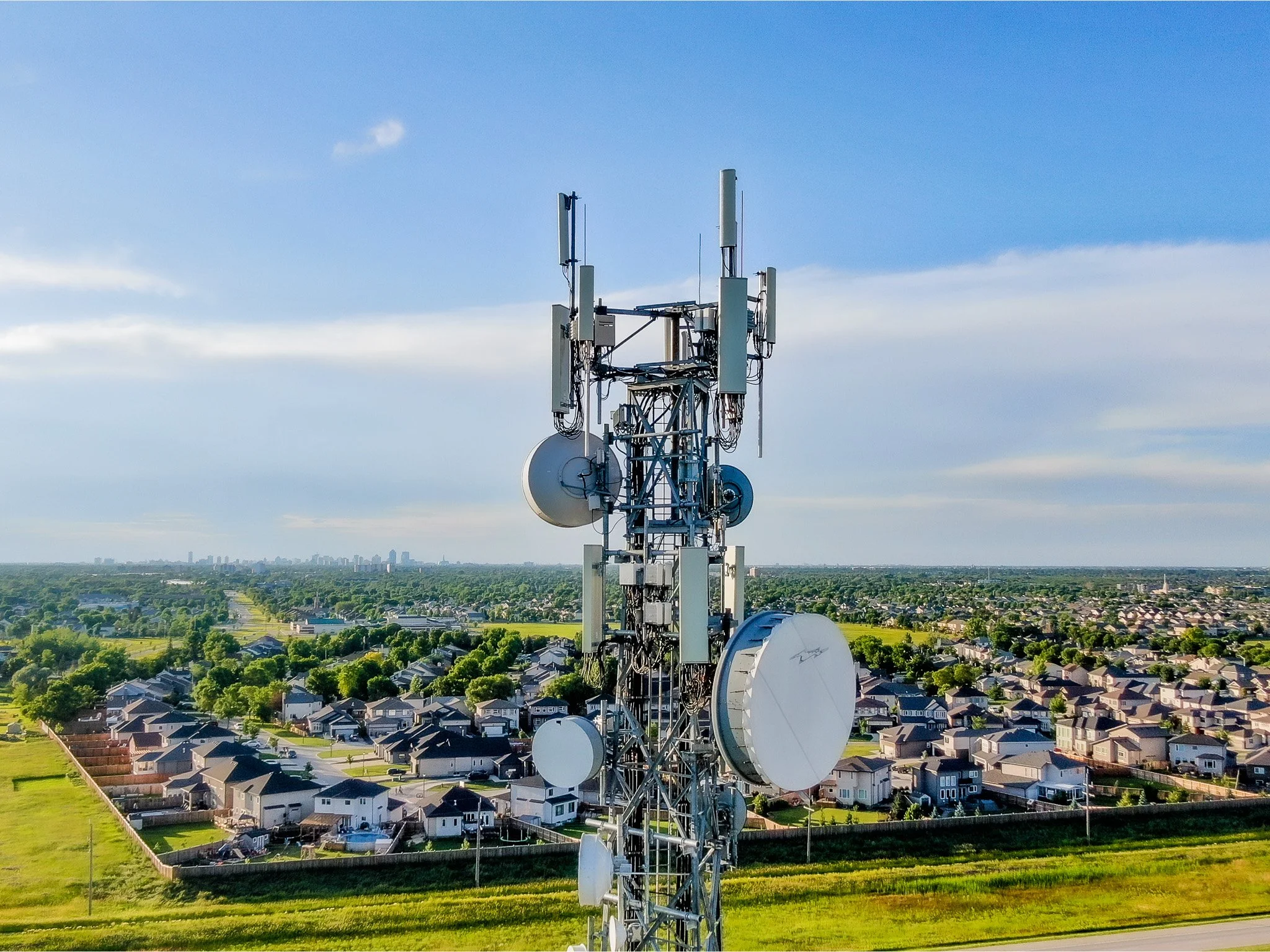 A tall telecommunications tower inspection with various antennas and dish receivers on top of a grassy hill overlooking a neighborhood of houses and a distant city skyline under a blue sky with some clouds Winnipeg Manitoba.