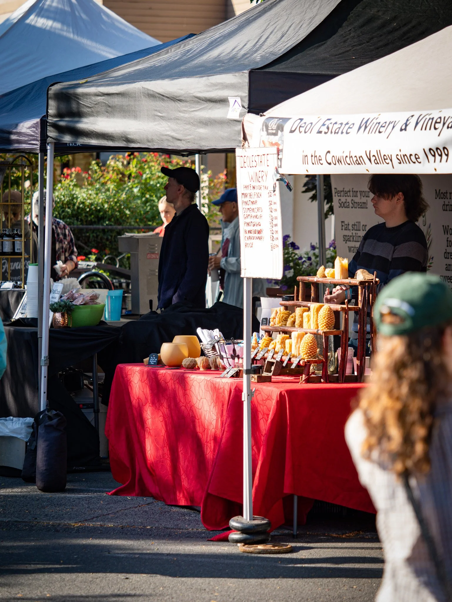 Market stall with red tablecloth selling candles and soaps, with a sign referencing Queen Elizabeth II, in an outdoor setting with people shopping Duncan BC Vancouver Island.