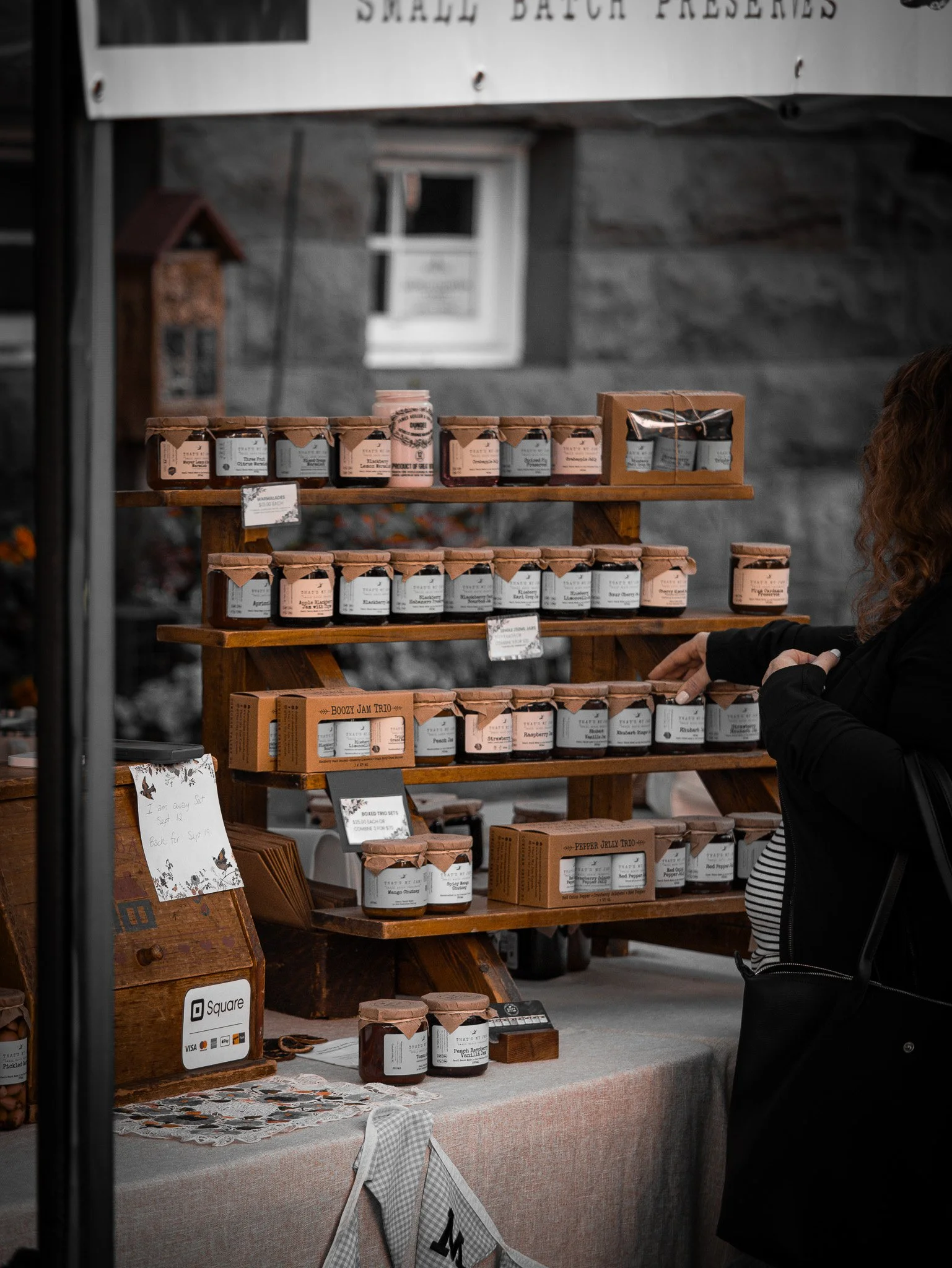 A market stall displays various jars of jam with labels, arranged on wooden shelves. A woman with curly hair is shopping, with only part of her arm and a striped shirt visible. There are signs and a Square payment sign on the table Duncan BC Vancouve