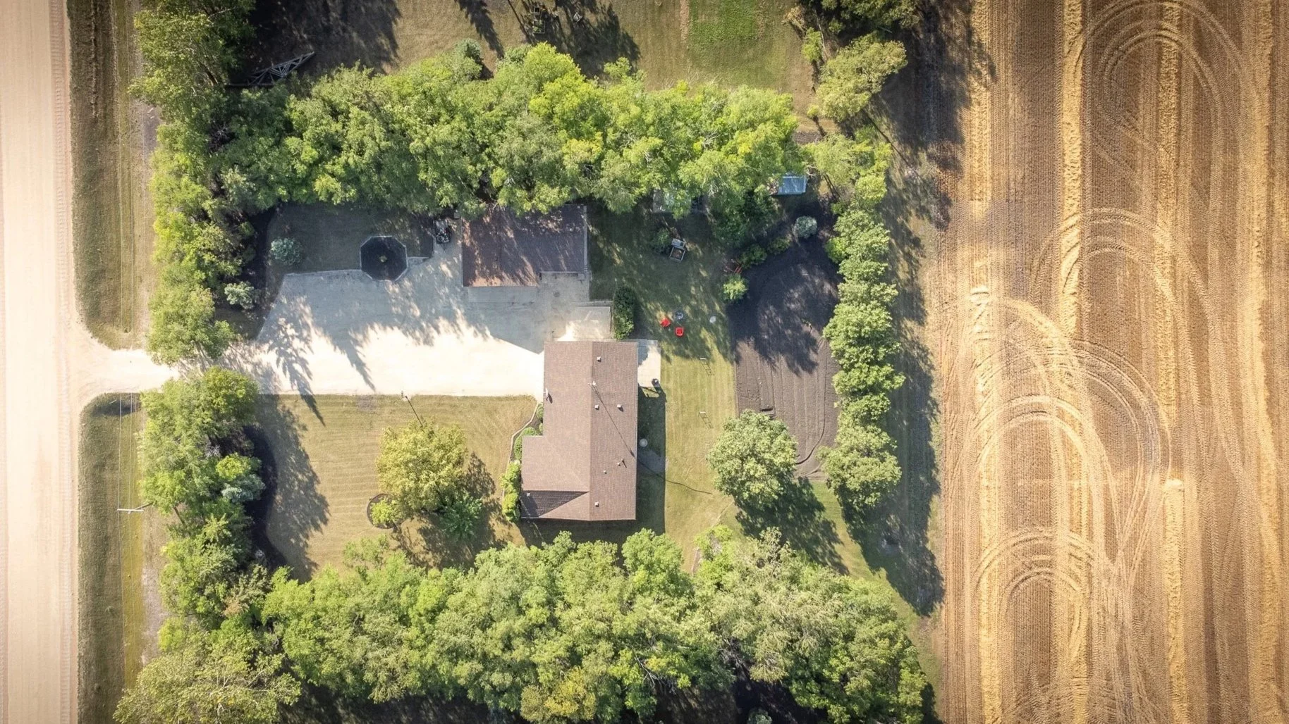 An aerial view of a house surrounded by trees, two sheds, a yard, and neighboring fields with a dirt road on the left side real estate photography Manitoba.