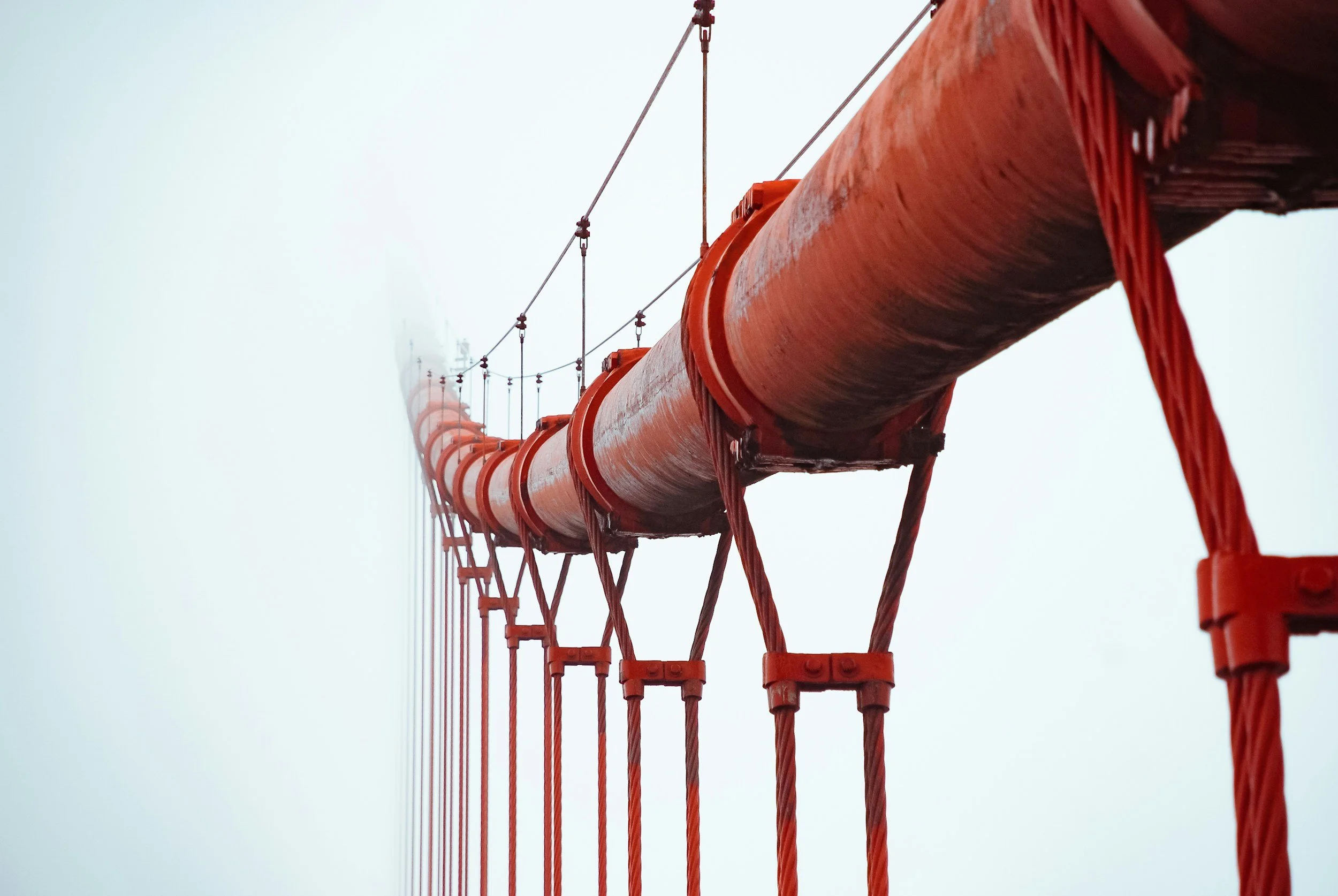 Large red suspension bridge cables and structural supports viewed from below against a pale sky