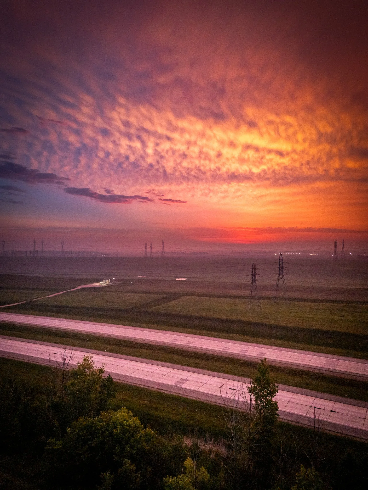 Colorful sunset sky with orange, pink, and purple clouds over green fields, highways, and power lines Winnipeg Manitoba.
