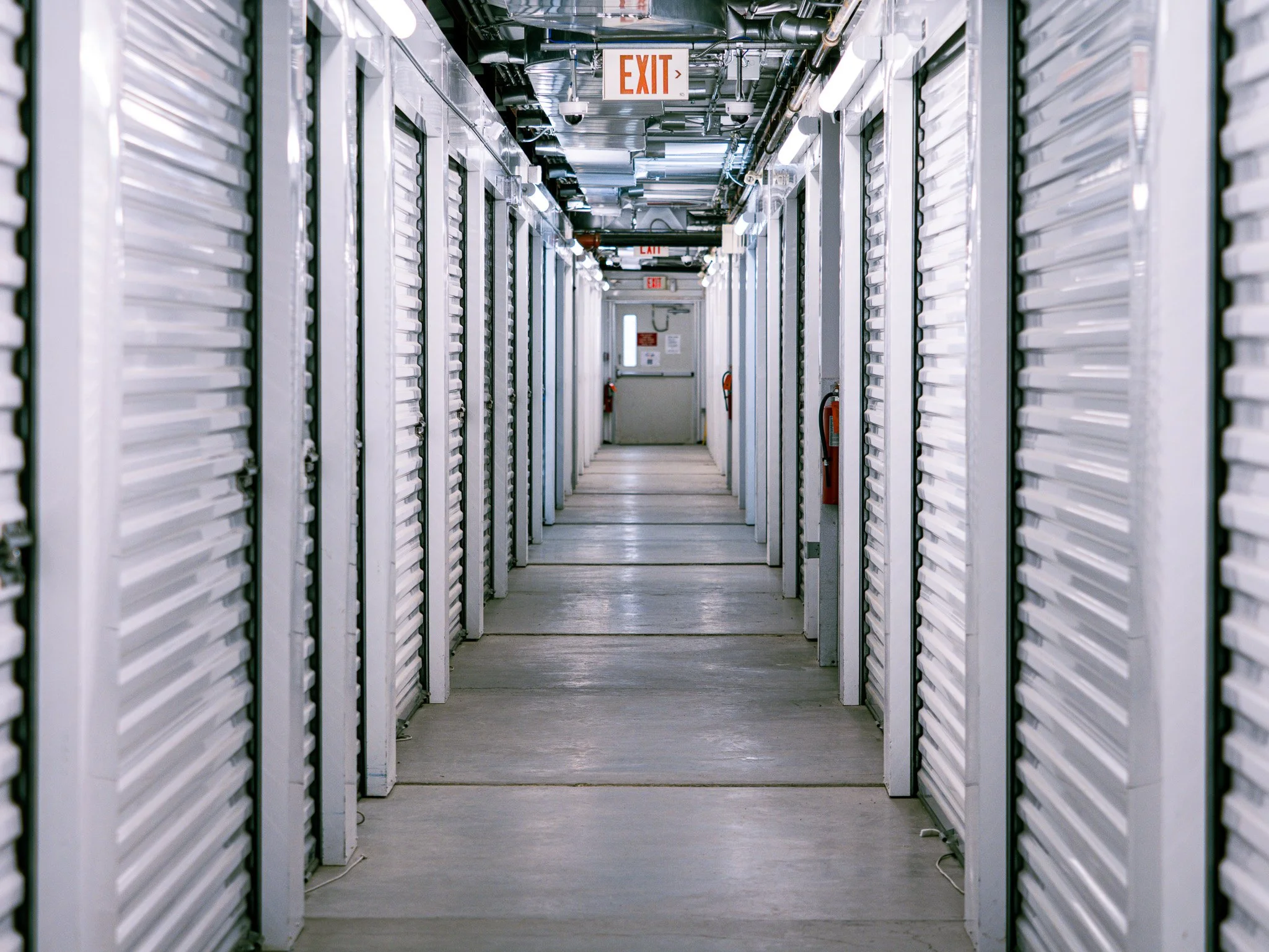 A corridor inside a data center with multiple server racks on both sides, fluorescent lighting, and an exit sign hanging from the ceiling.