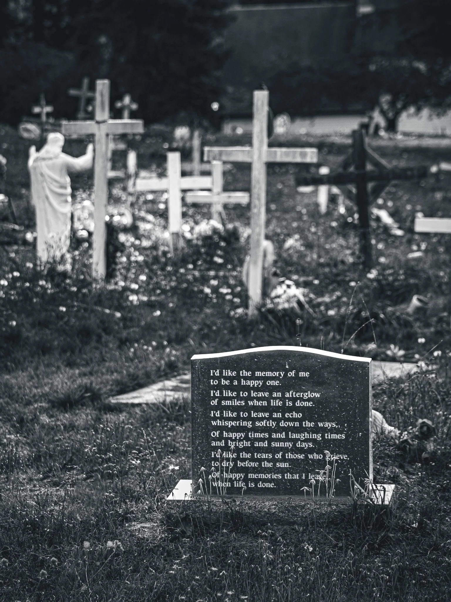A black and white photograph of a cemetery with a headstone in the foreground containing a poem about memories and happiness. In the background, there are several crosses and a statue of Jesus Christ with outstretched arms among the graves. The scene