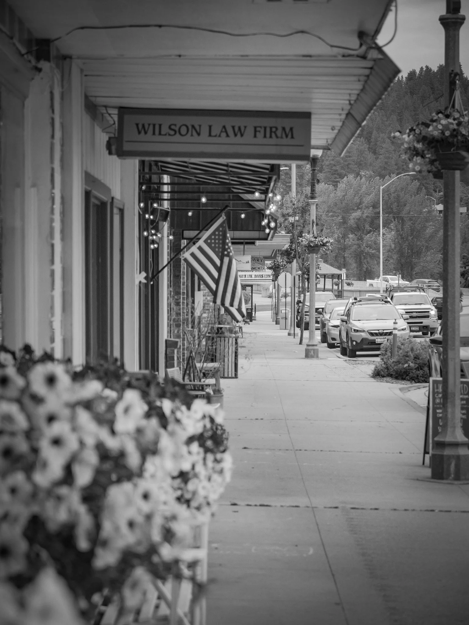 Black and white photo of a sidewalk in a small town, with parked cars, streetlights, hanging flower baskets, and a sign for Wilson Law Firm