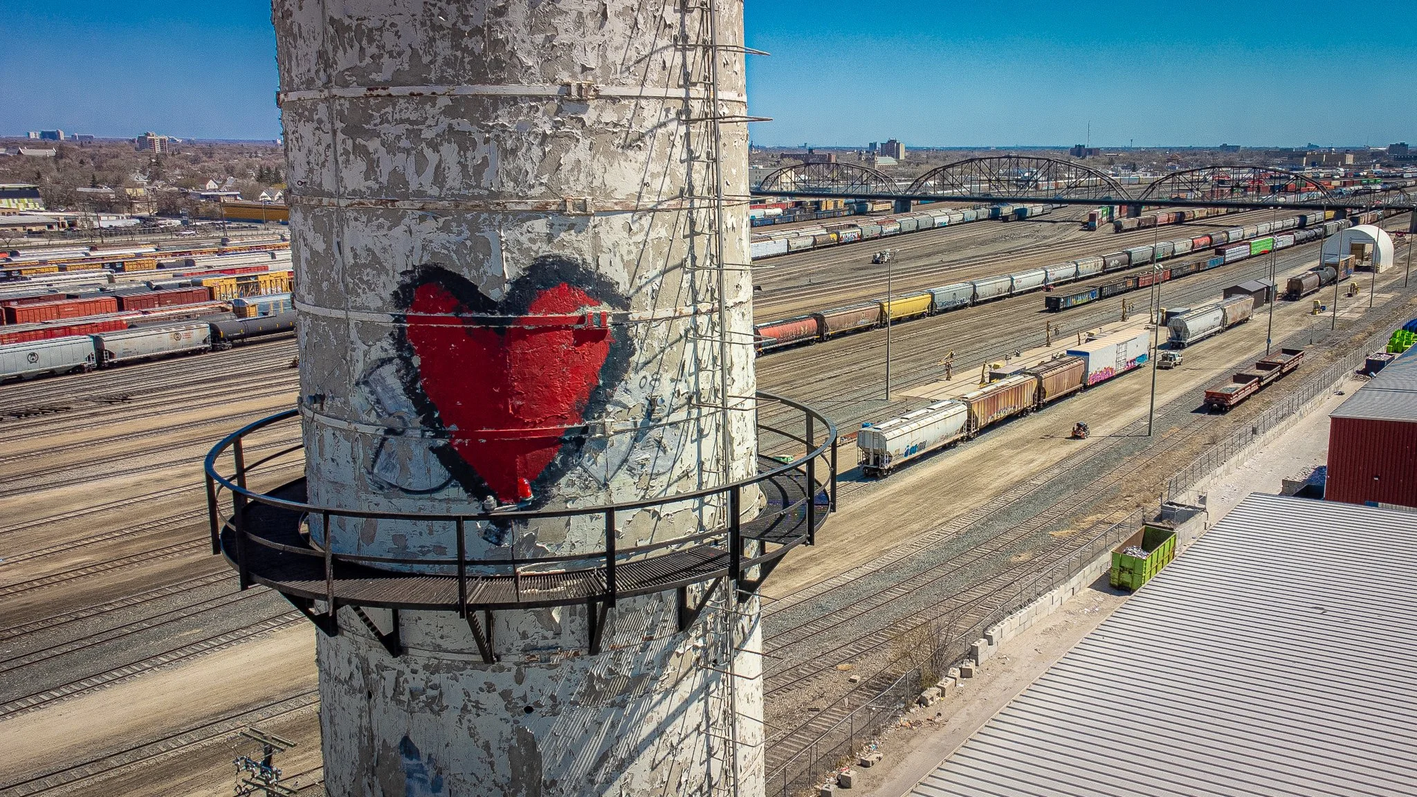 Rail yard filled with freight trains, viewed from a weathered concrete tower painted with a large red heart, with arched bridges in the background. winnipeg aerial roof and building envelope inspections service