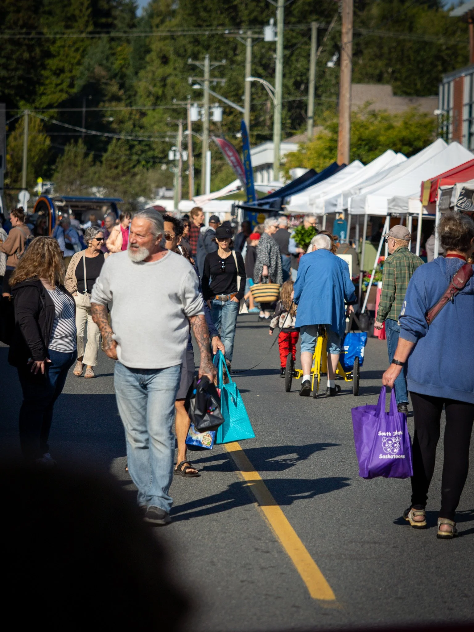 People walking and shopping at an outdoor street market with white canopy tents on a sunny day, green trees in the background, and power lines overheadDuncan BC Vancouver Island.