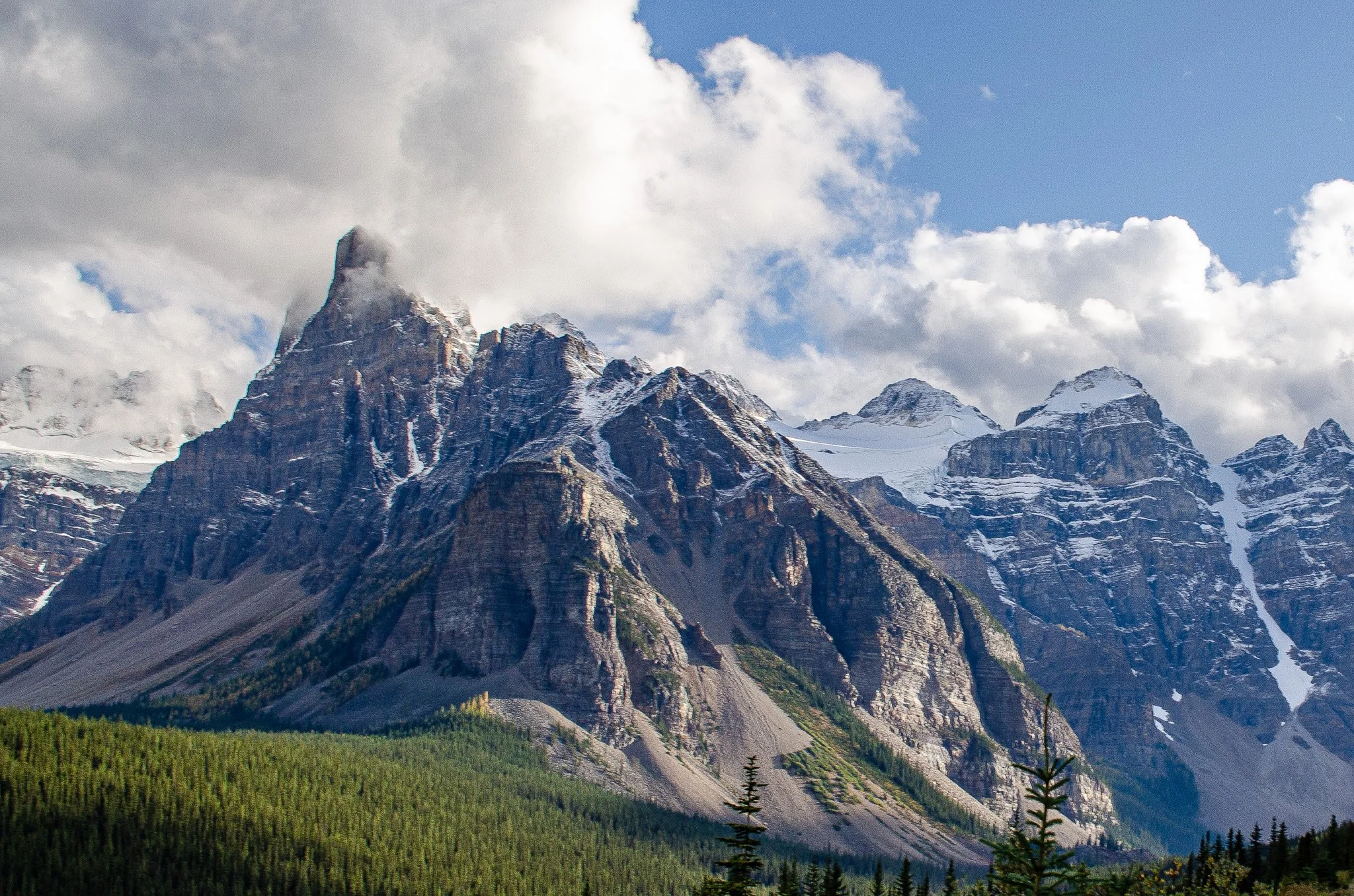 Snow-capped mountain peaks with partially cloudy sky and green forested valley below Canmore Alberta