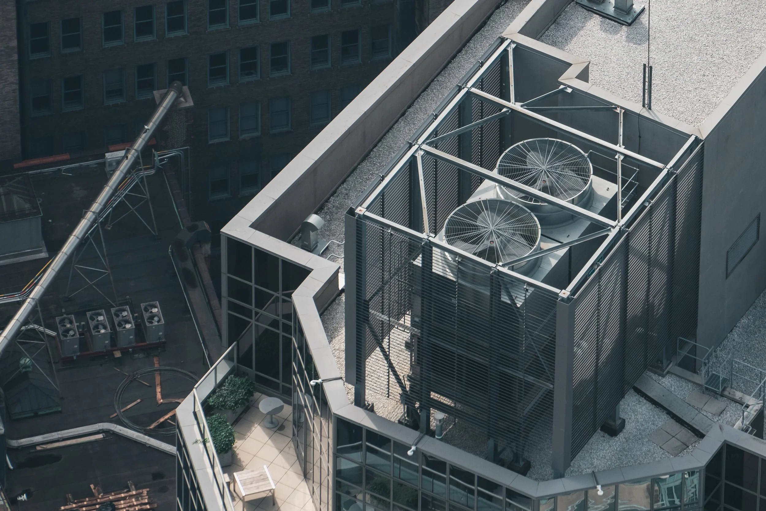 A rooftop view of a modern office building showing HVAC units, air vents, and a small rooftop patio with chairs and potted plants.