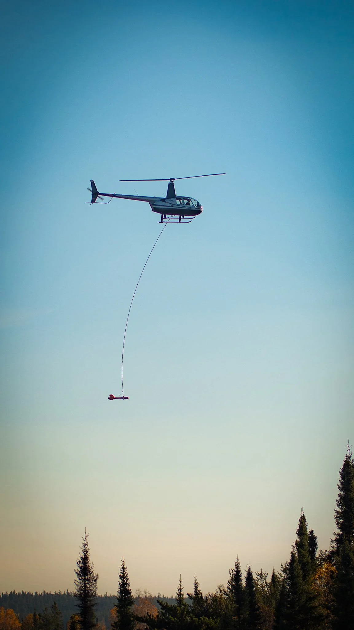 A helicopter flying in the sky, towing an object connected by a cable, with a forest at the bottom Fox Mine Manitoba 