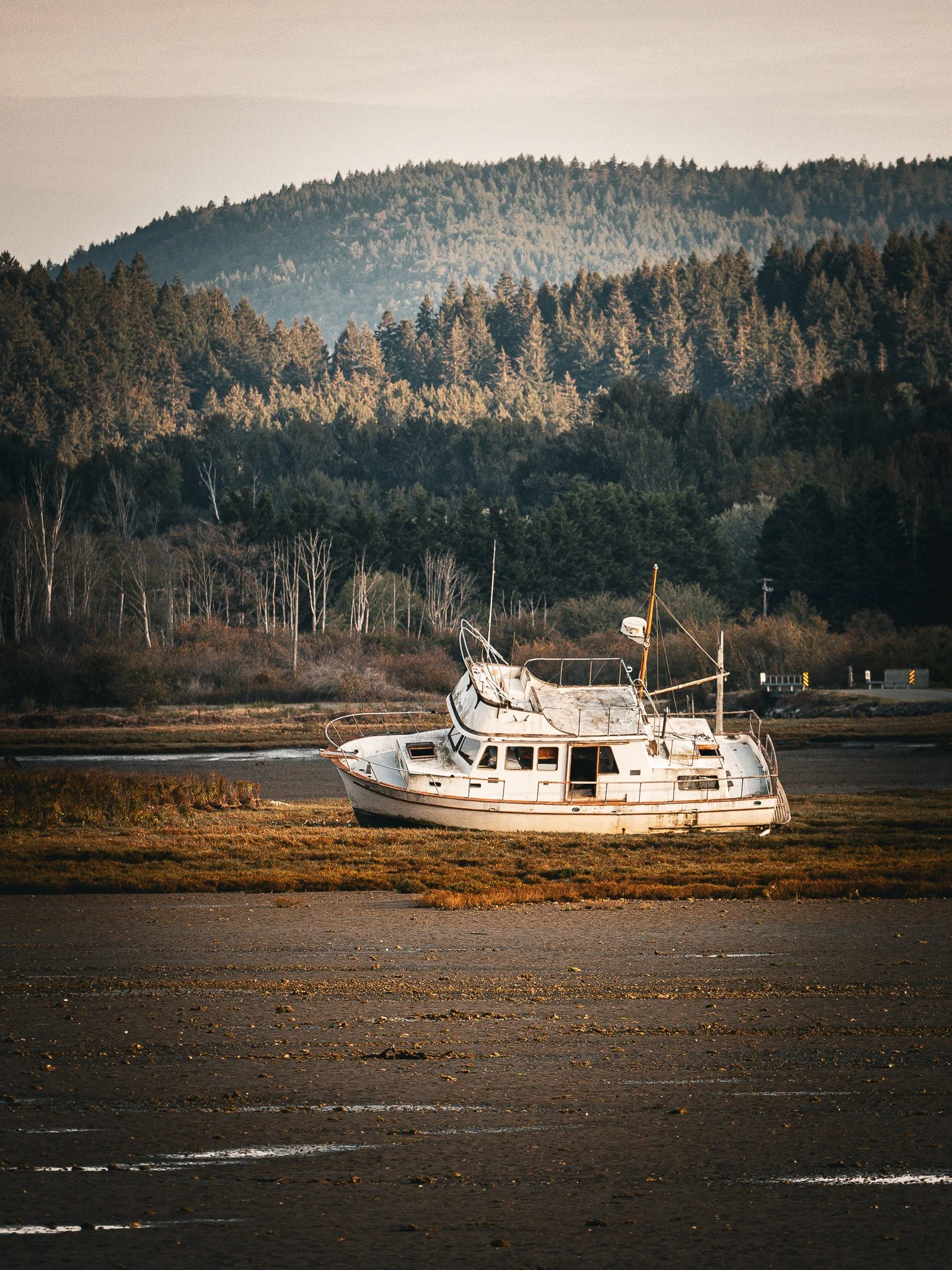 An abandoned, weathered boat on a dry, cracked ground near a forested hillside and mountain range in the background during sunset Cowichan Bay Vancouver Island.