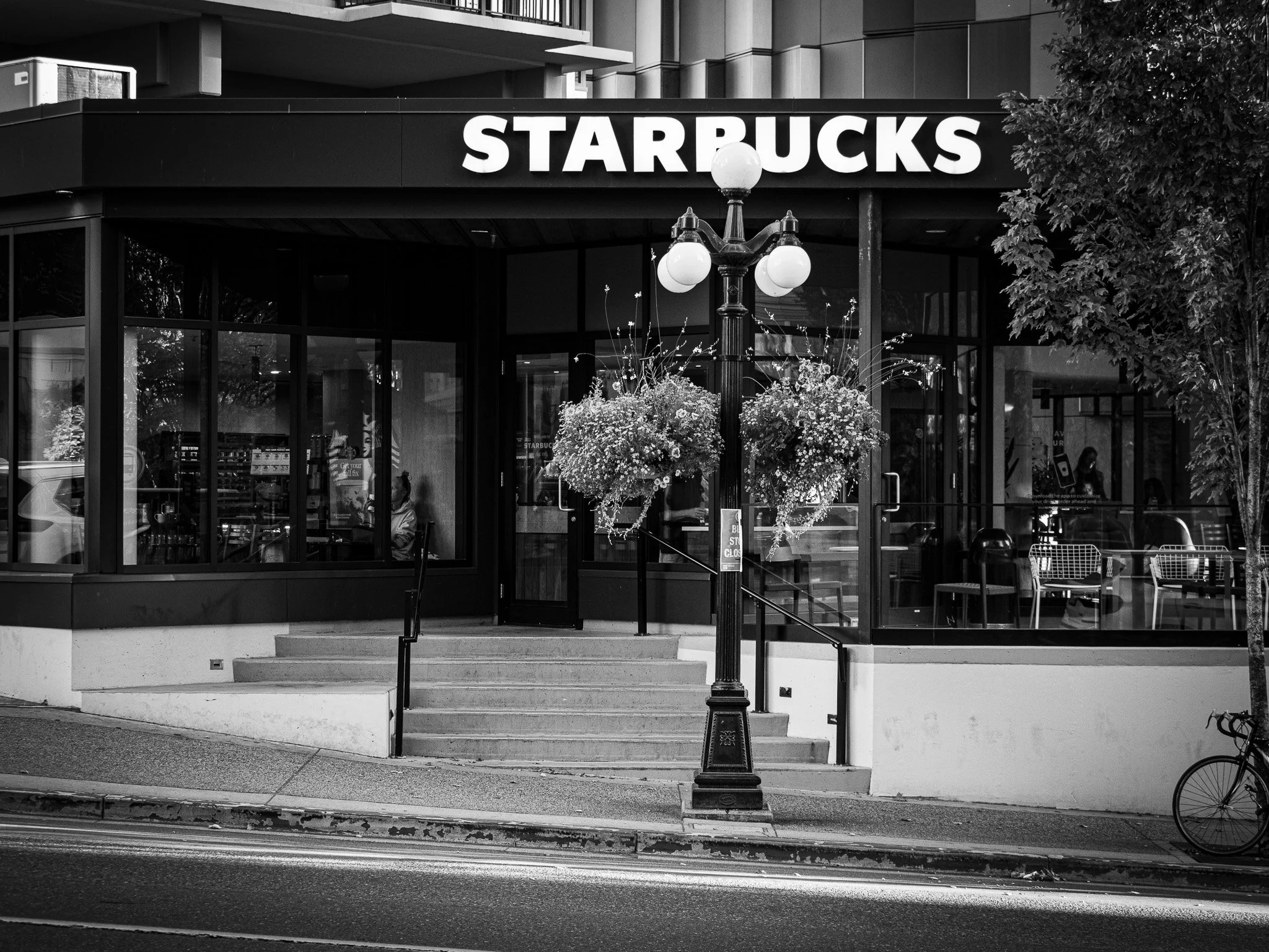 Starbucks streetscape: Black and white street photograph of a Starbucks storefront, framed by a decorative street lamp with hanging flower baskets and a bicycle parked nearby Victoria BC
