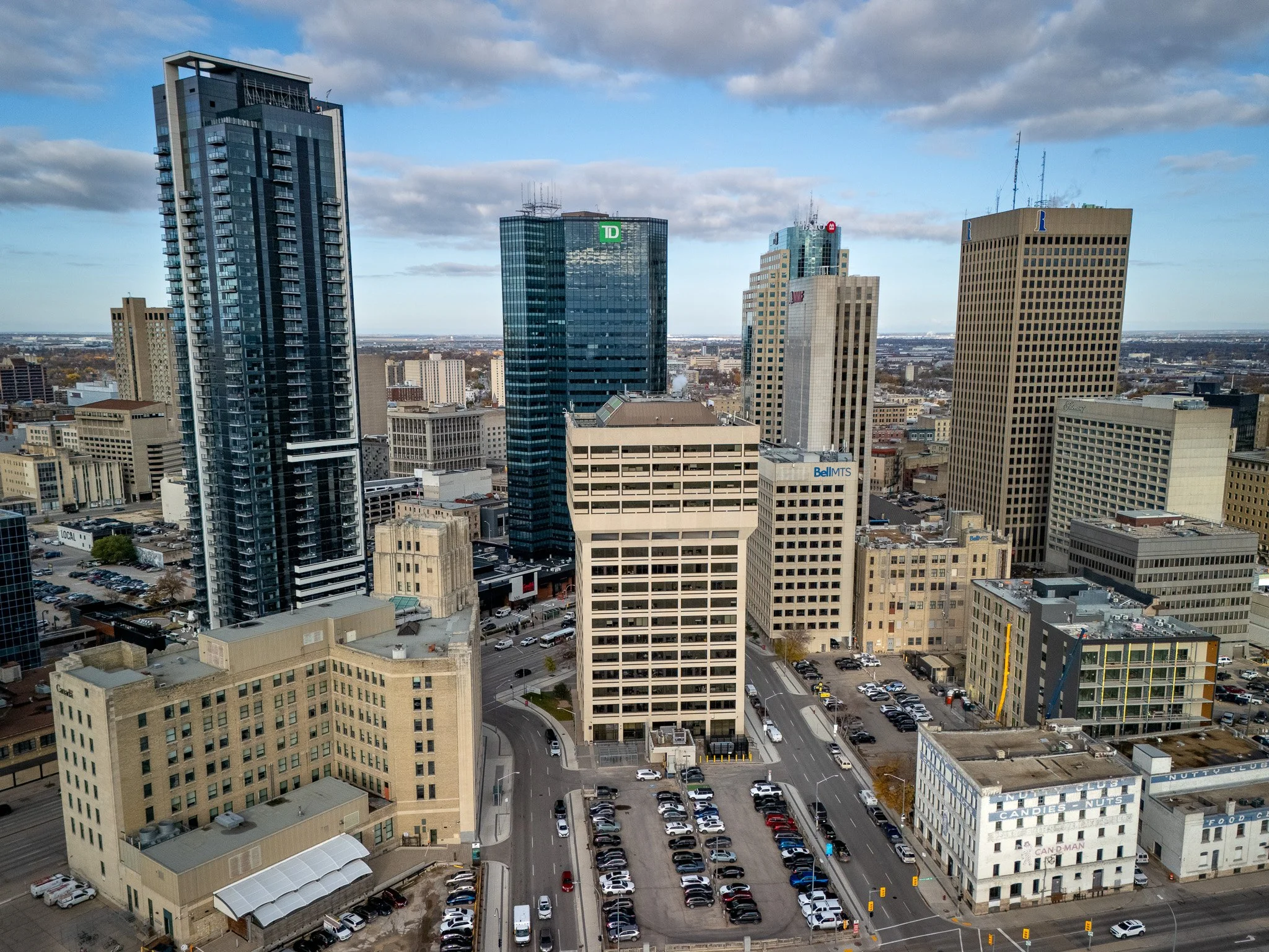 A city skyline with tall modern skyscrapers, including a building with the TD logo and a building with a red 'M' logo, under a partly cloudy sky. Streets are filled with parked cars and some moving vehicles. Winnipeg Skyline aerial photo