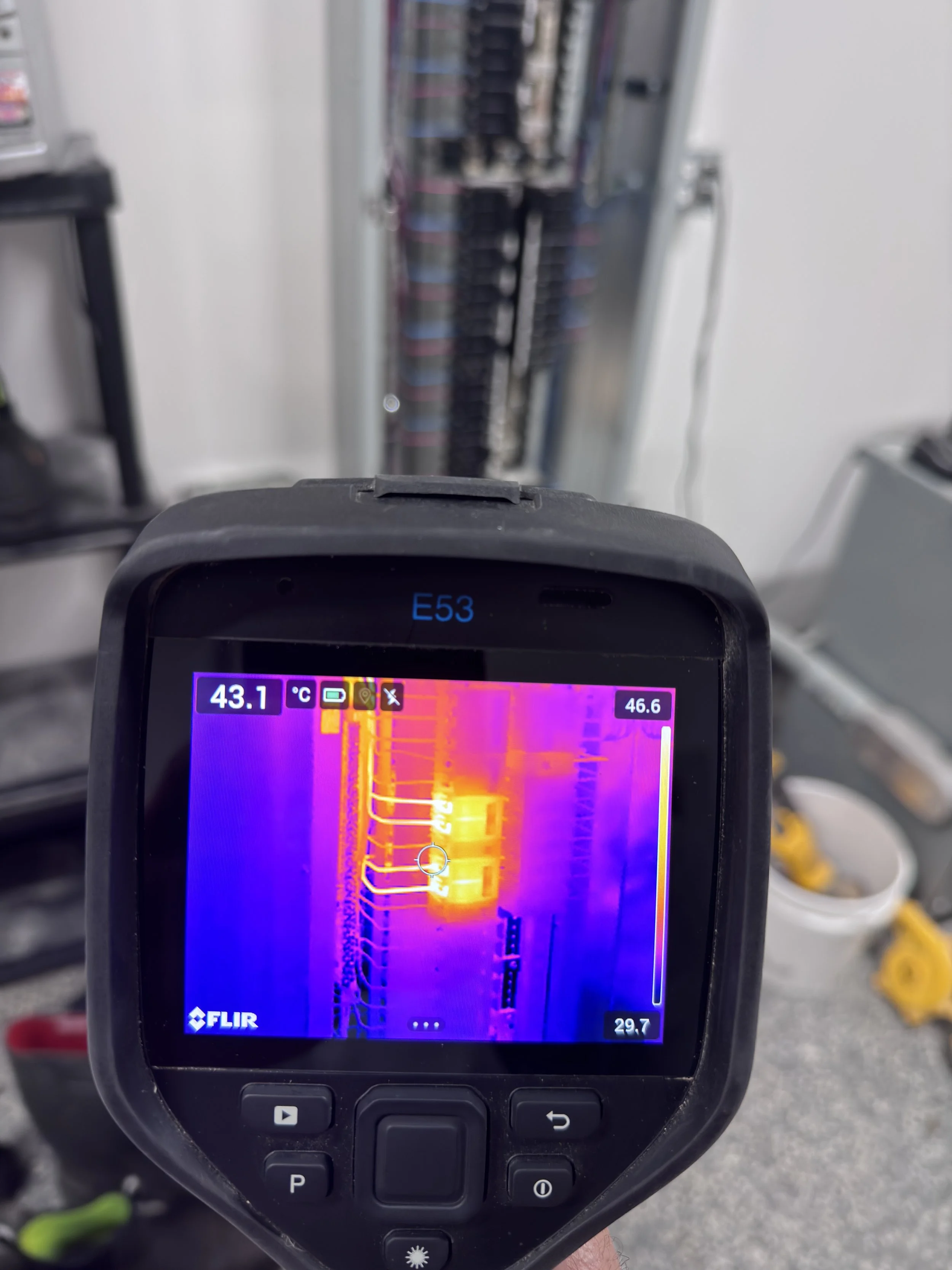 Thermal imaging device displaying temperature readings in a workshop with a fuse box in the background.