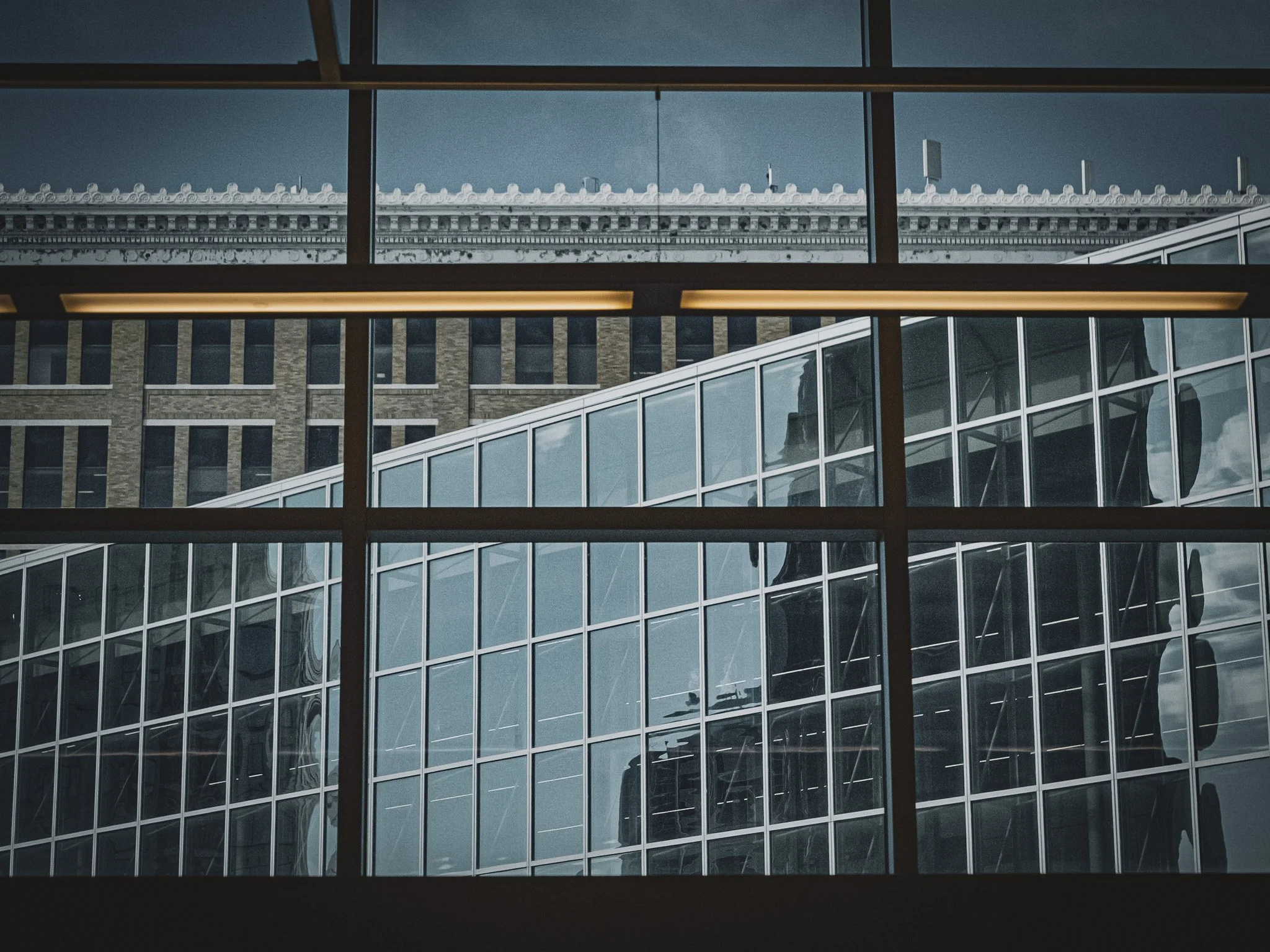 View of modern architectural building through large window panes with visible reflections, including a rooftop with decorative cornice details and a cloudy sky.