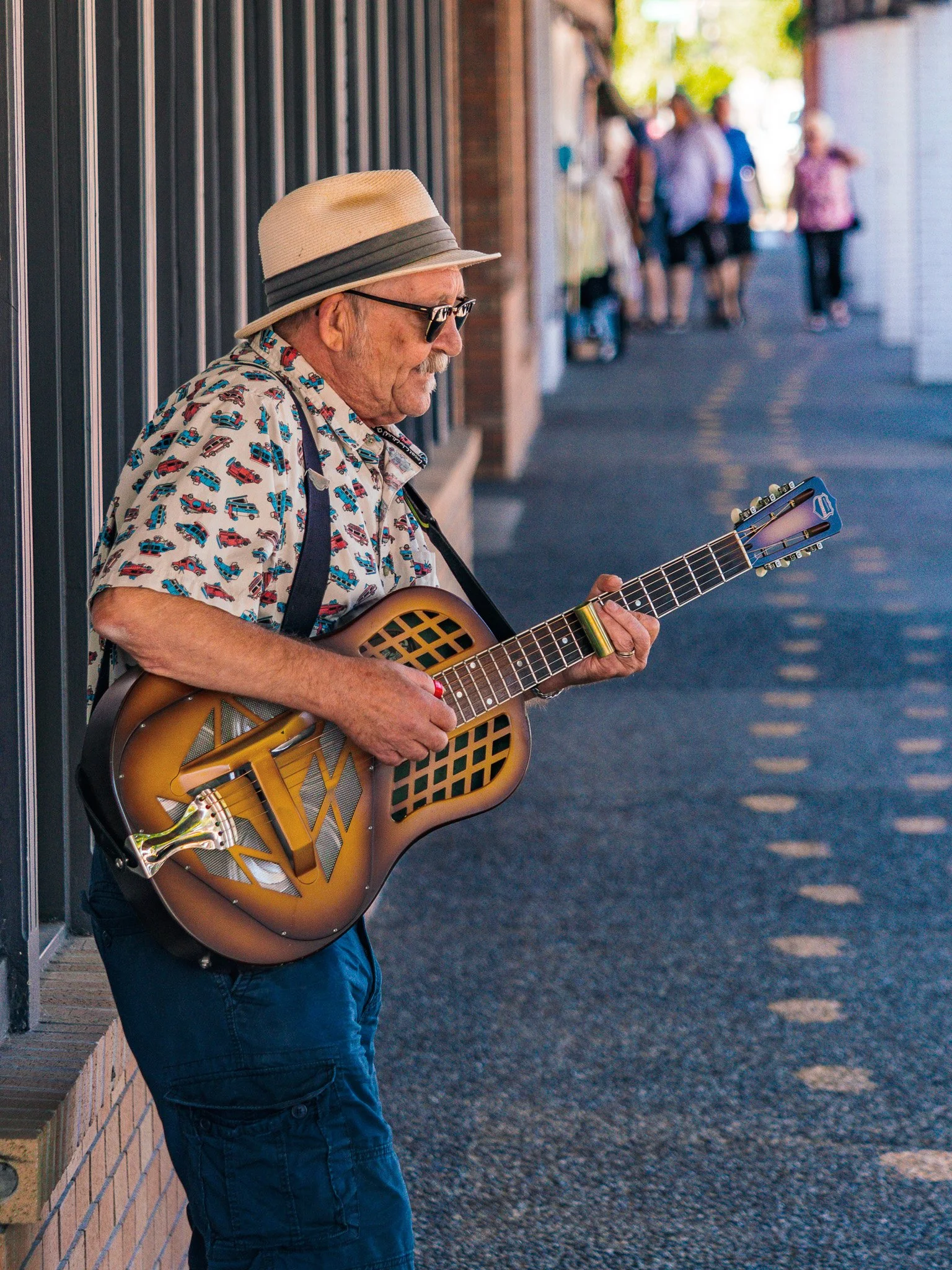 An elderly man wearing a straw hat, sunglasses, and a colorful short-sleeved shirt with cars printed on it, playing an acoustic guitar while leaning against a brick wall on a street, with a group of people walking Chemainus BC
