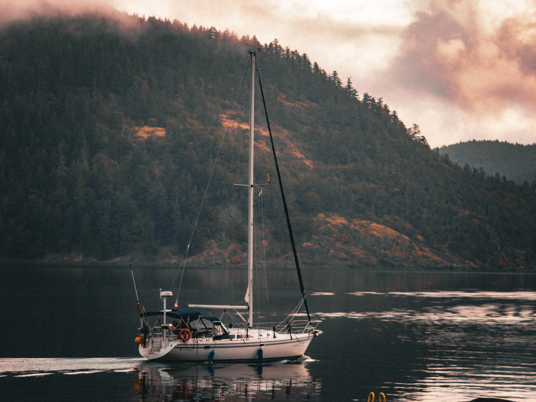 A sailboat on calm water with a mountainous landscape in the background, partly covered with fog and clouds, during sunset or sunrise Cowichan Bay Vancouver Island.