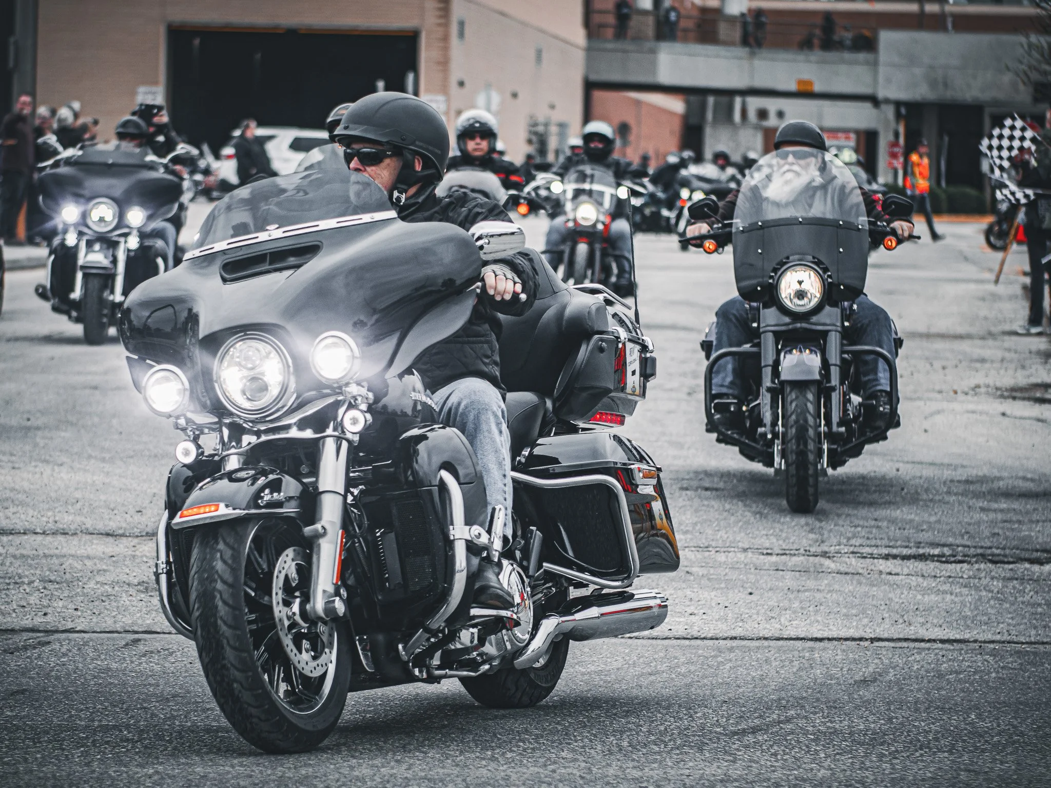 A group of motorcyclists wearing black helmets and jackets riding on a city street, with buildings and onlookers in the background ride for dad Winnipeg MB.