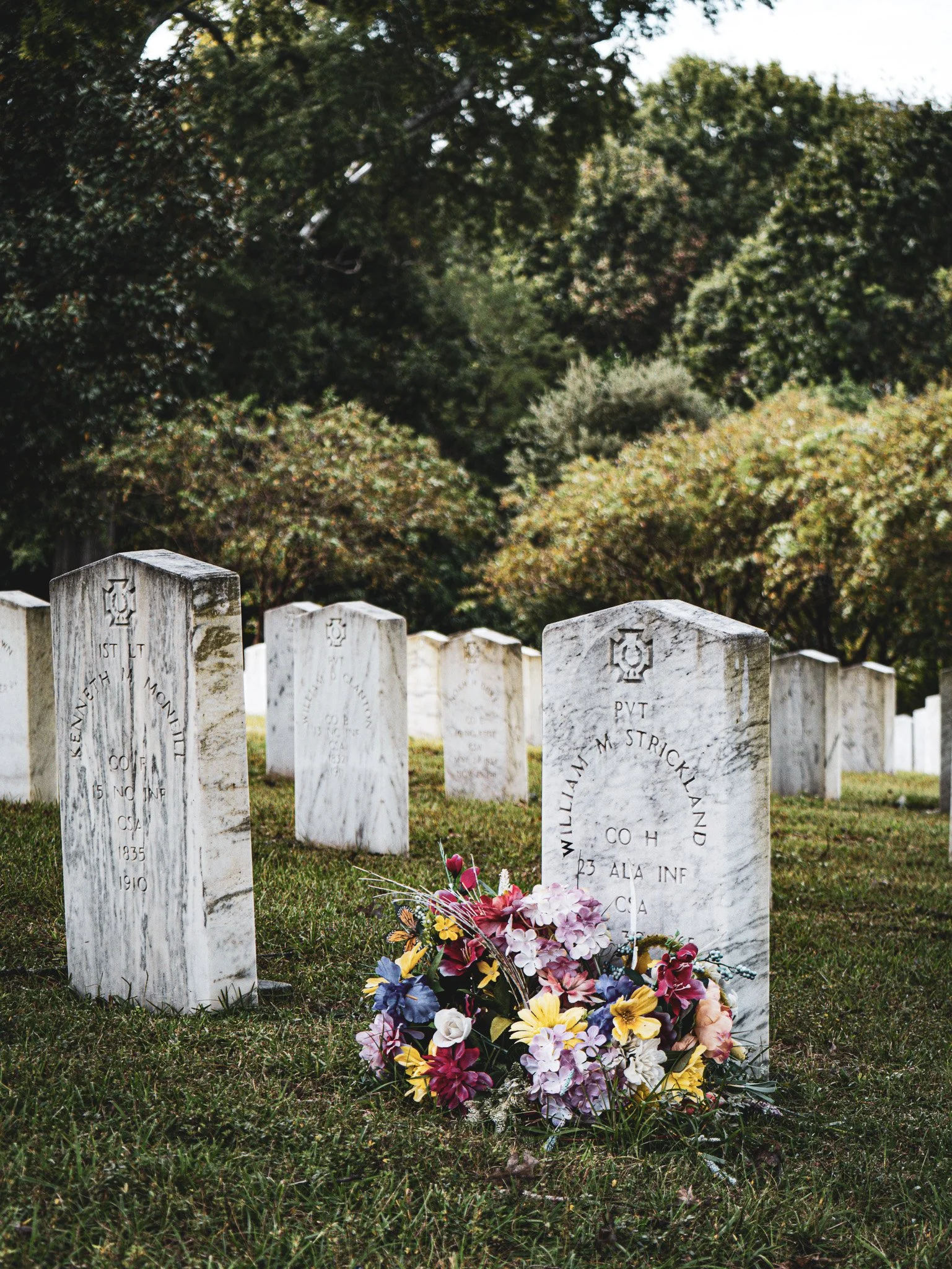A graveyard with white marble headstones and colorful flowers at the base of one marker, with green trees in the background North Carolina military graveyard unknown soldier.