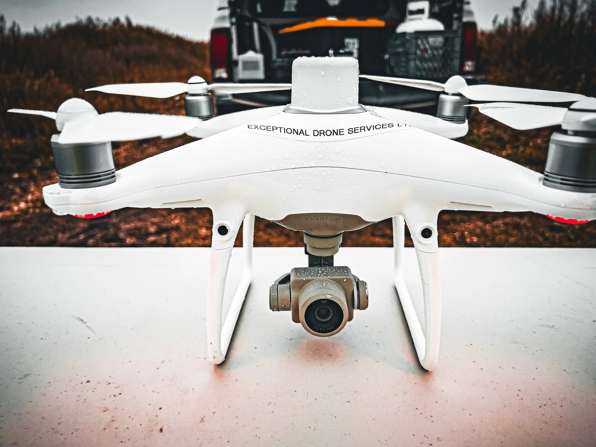 A white drone with multiple rotors and a built-in camera on the bottom, labeled 'EXCEPTIONAL DRONE SERVICES LTD,' sitting on a white surface with a black vehicle in the background. The drone's propellers are visible, and the background appears to be 