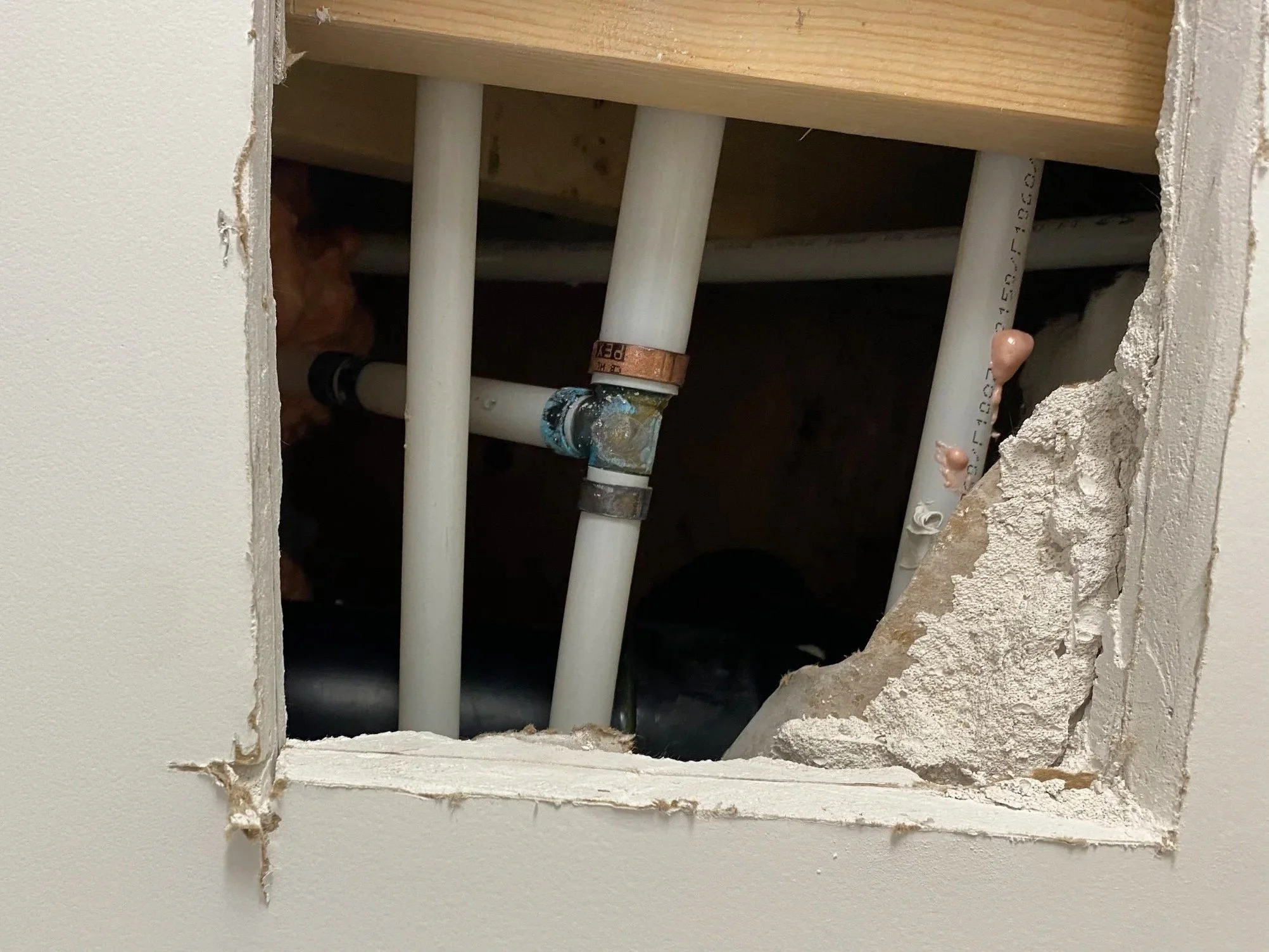 View of plumbing pipes behind a section of drywall cut out, with some parts showing joints, and a small orange piece attached to one pipe, in a construction or repair setting.