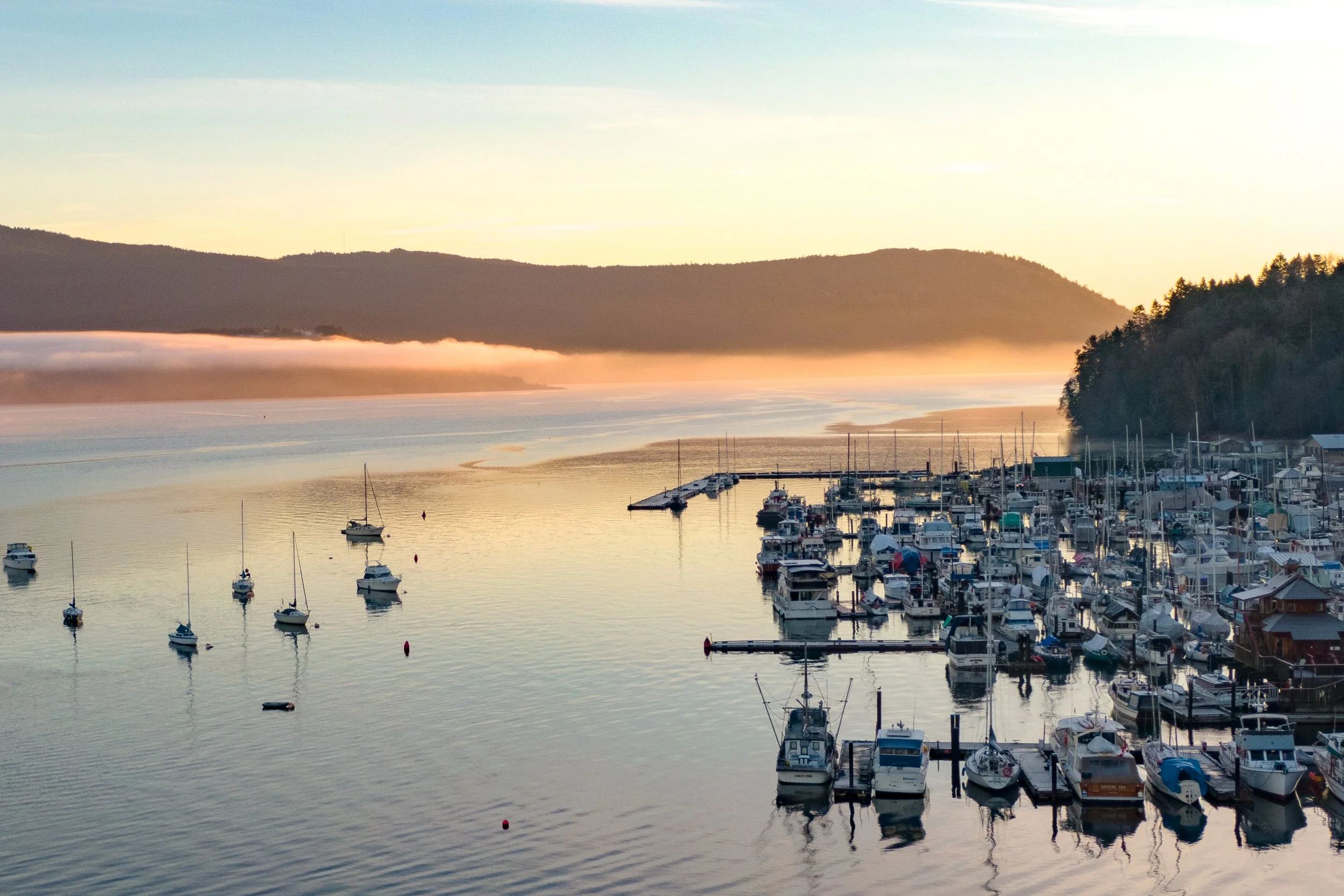 A marina with boats docked along a pier, calm water reflecting the sky, forested hills in the background, and the sun rising or setting casting a golden glow.
