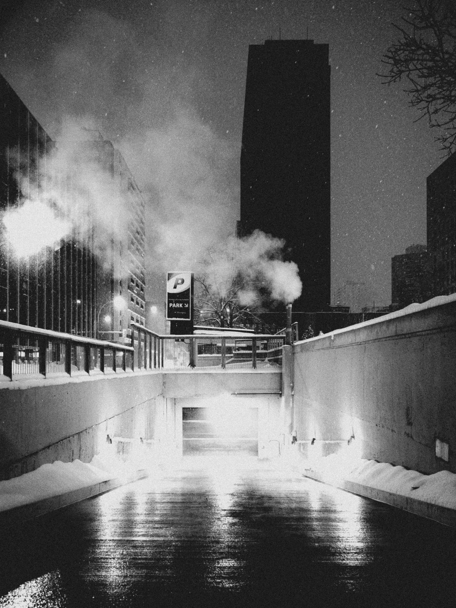 Black and white photo of a snowy urban scene at night, showing a tunnel or underpass, with buildings, smoke, and a parking sign in the background Winnipeg Manitoba.
