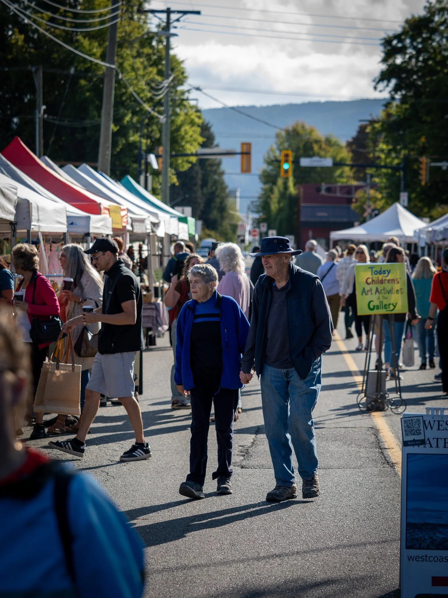 Crowd of people walking and standing at an outdoor market with tents, trees, and mountains in the background Duncan BC Vancouver Island.
