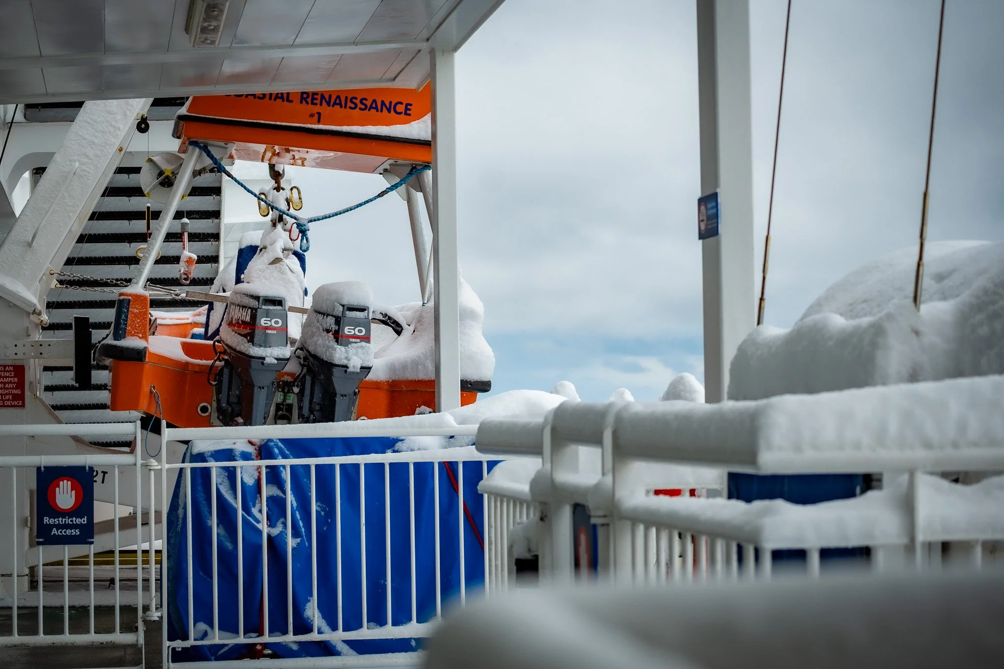 Lifeboats labeled as 'Coastal Renaissance #1' covered in snow, on a ship deck, with safety bars and restricted access signs, foggy sky in the background Nanaimo Vancouver Island.